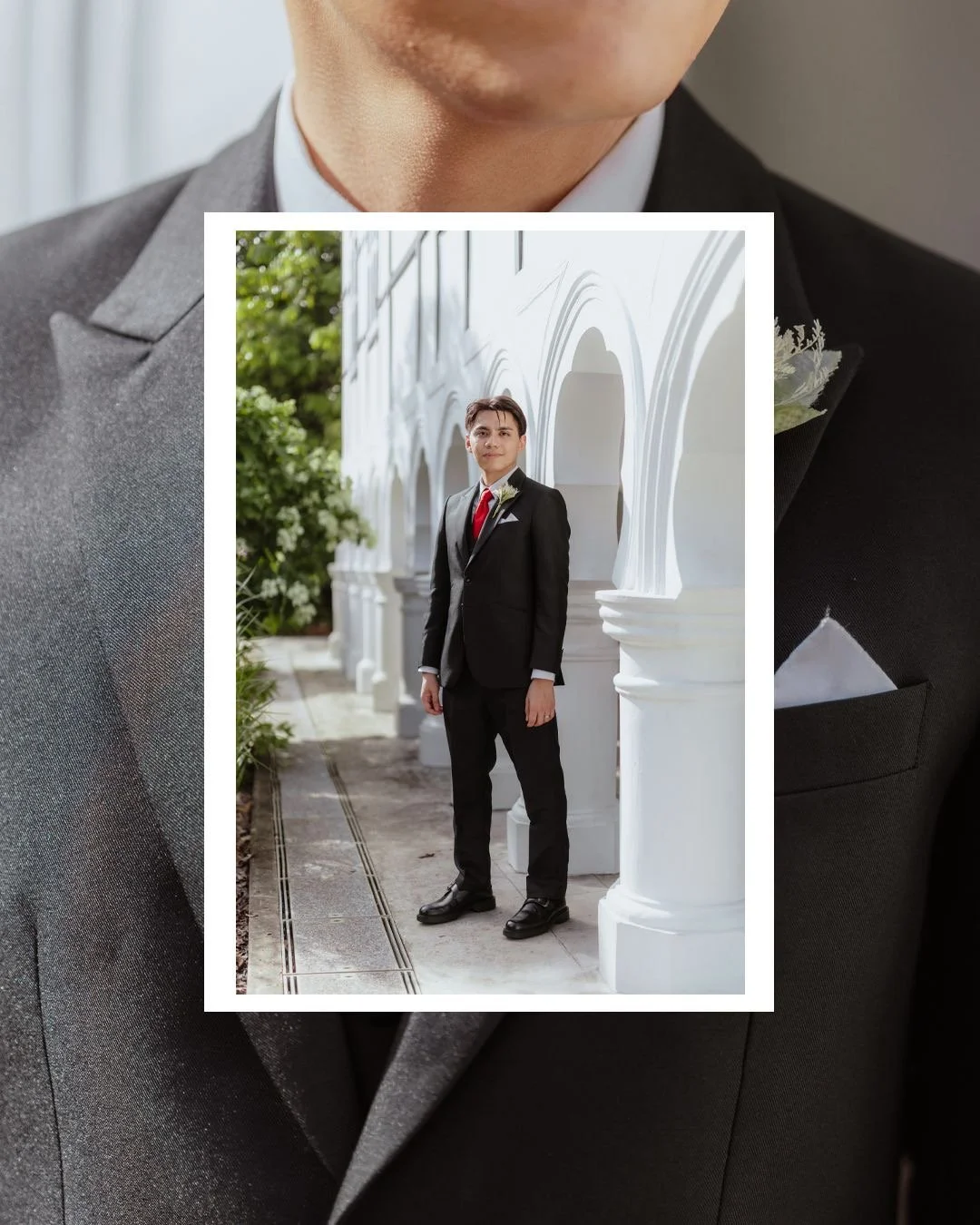 A man in a black suit with a red tie and boutonniere standing outside near white architectural columns and greenery.