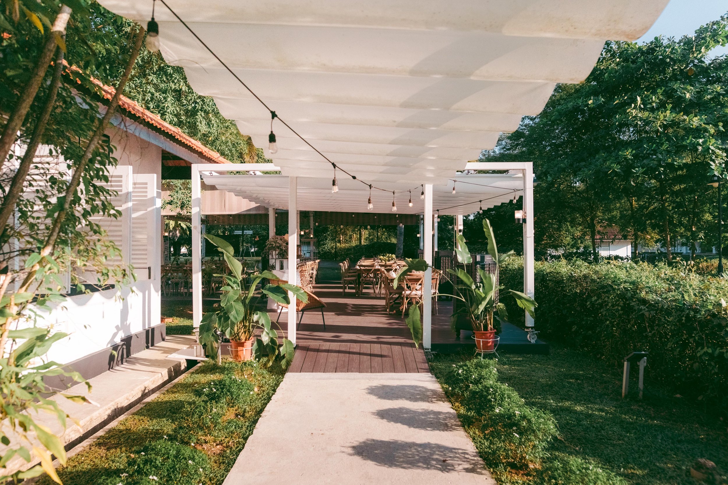 Outdoor patio with tables and chairs, surrounded by greenery and potted plants, with a white pergola and string lights overhead.