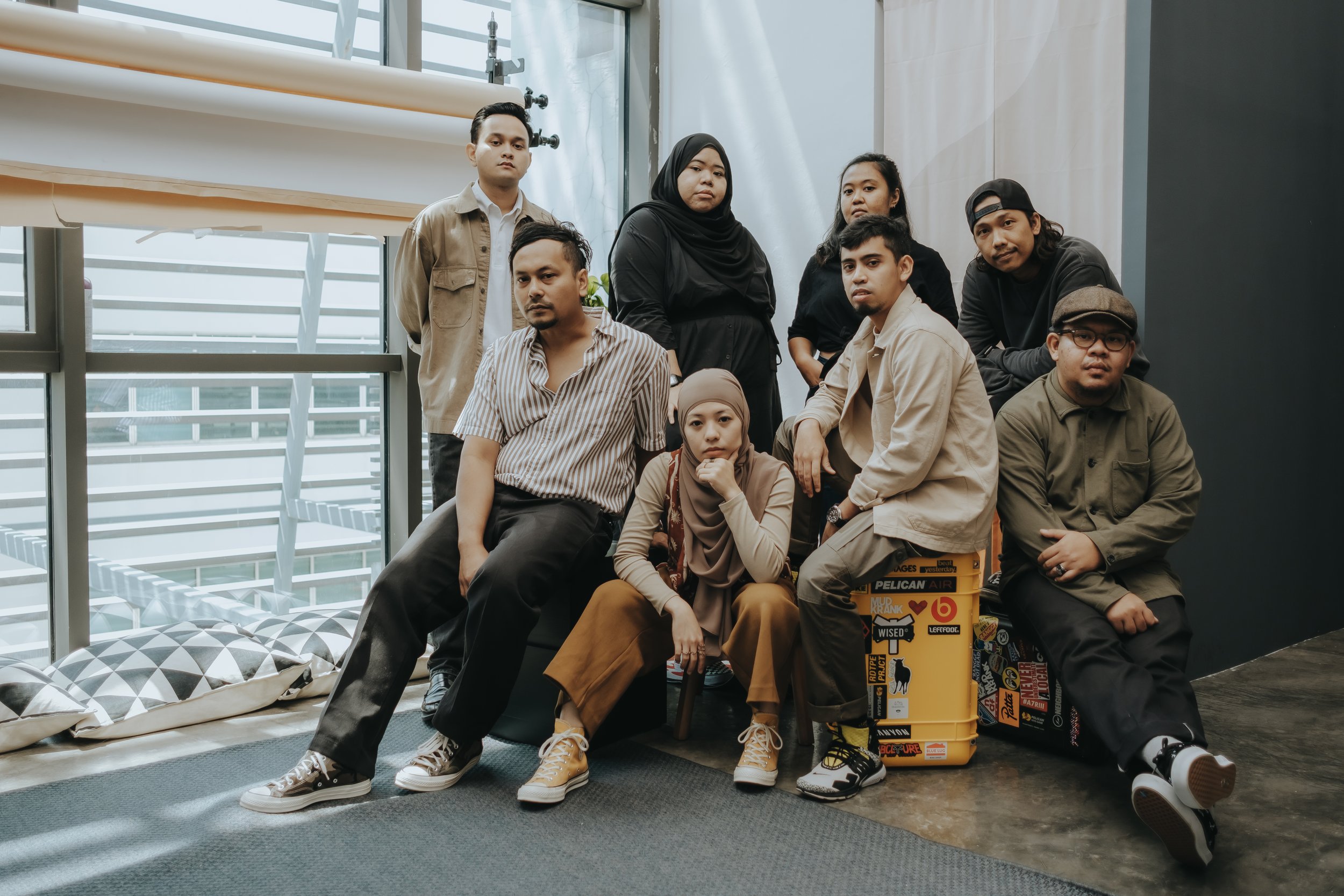 A group of nine diverse young adults, both men and women, posing indoors against a modern, glass-walled backdrop with natural light. Some are sitting on the floor, others standing behind, dressed in casual, trendy clothing.