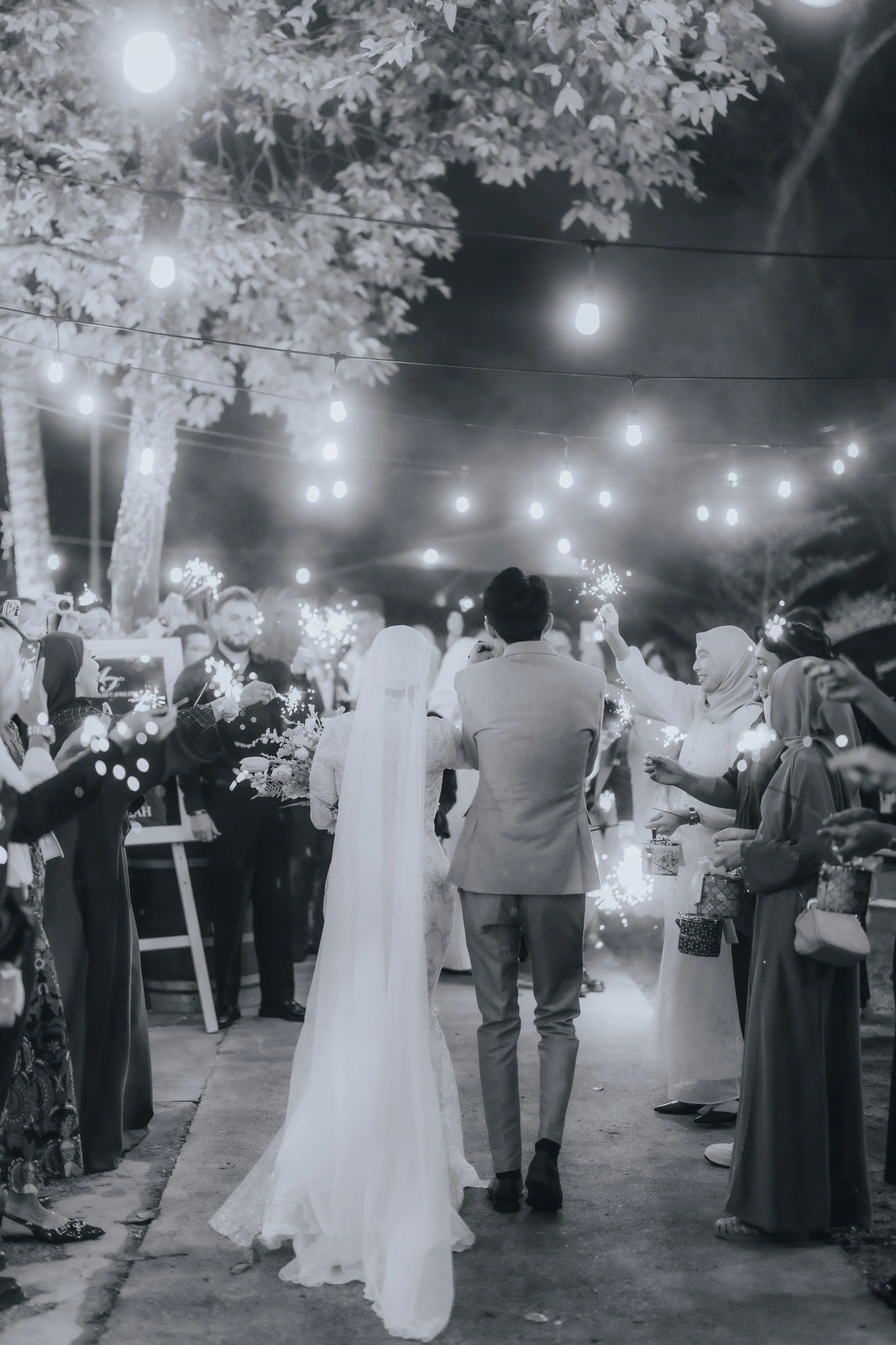 A black and white photo of a wedding celebration at night, with a bride and groom walking away from the camera, surrounded by guests holding sparklers, under string lights and trees.