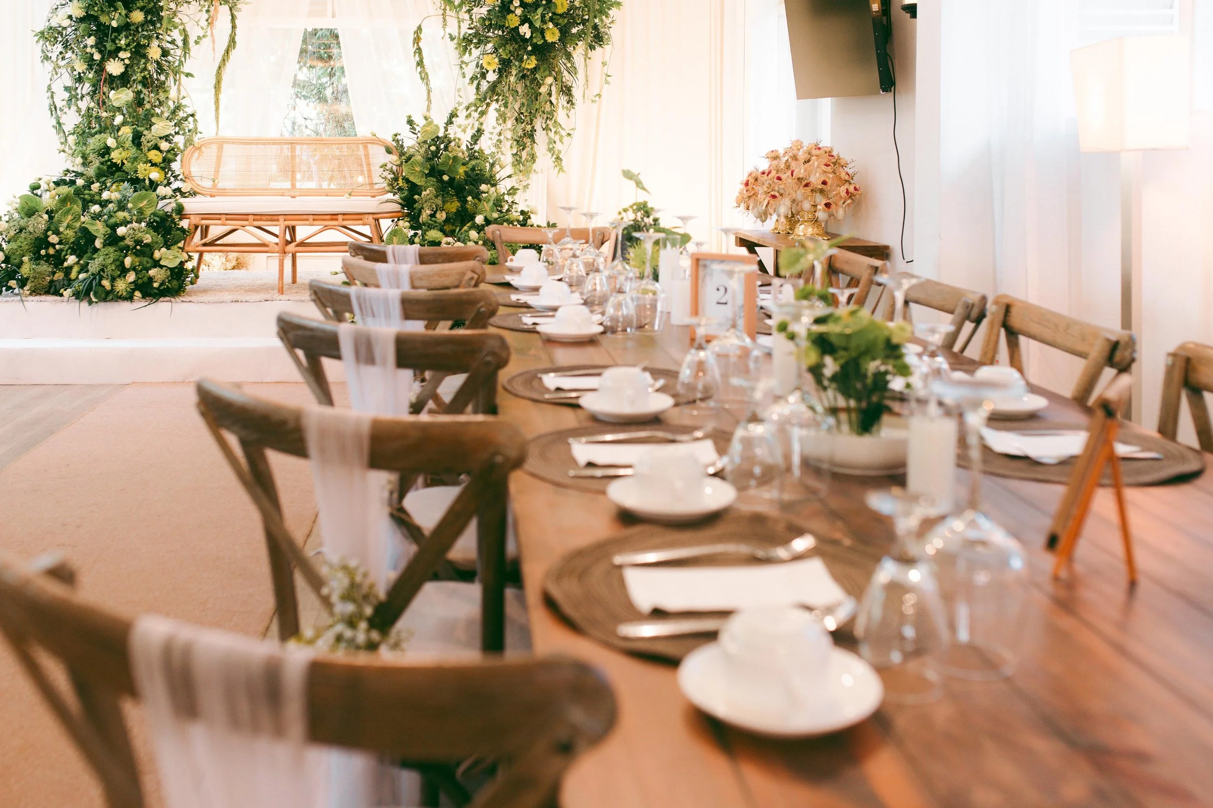 A long wooden dining table decorated with white dishes, glasses, cutlery, and floral centerpieces, set for a wedding reception in a bright room with white curtains and a floral backdrop at the head of the table.