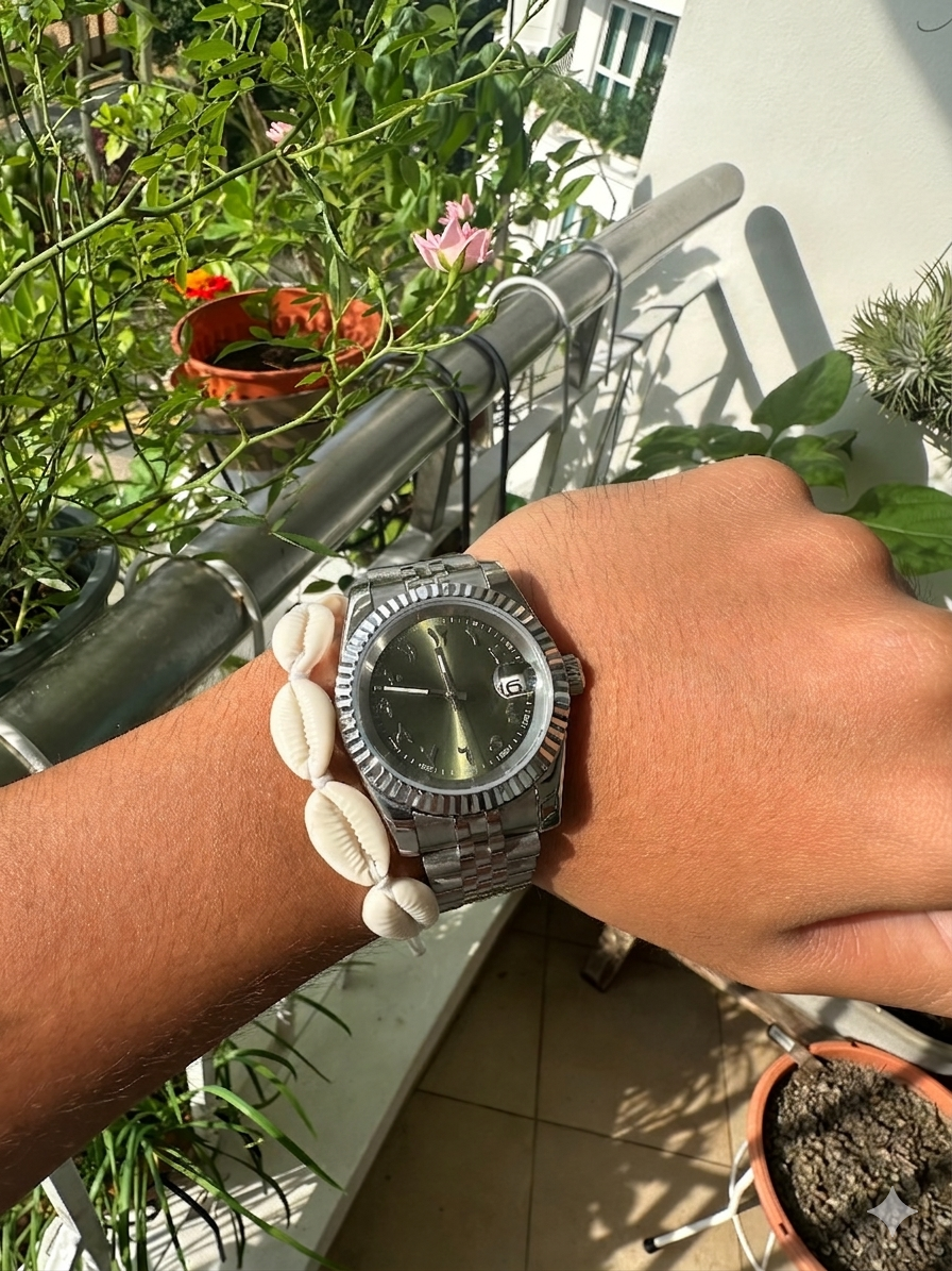 A person's wrist wearing a silver wristwatch with a dark face, alongside a shell bracelet, with a balcony filled with potted plants and flowers in the background.