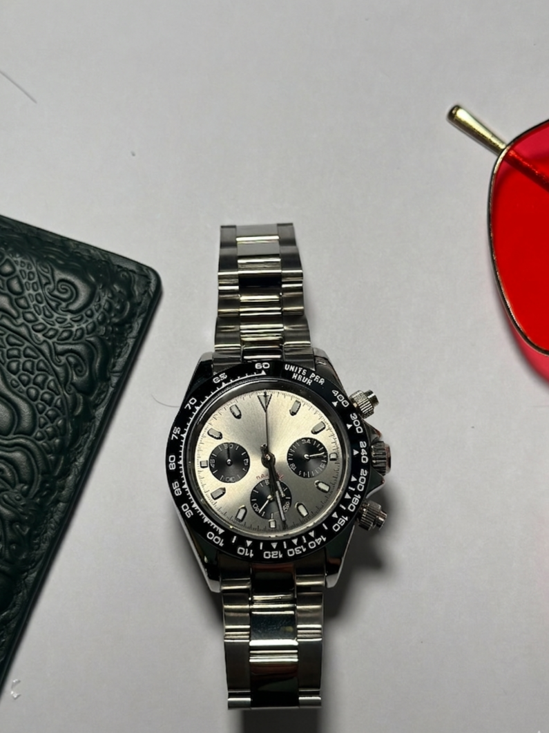 A silver wristwatch with a black bezel and three subdials resting on a white surface, partially surrounded by black textured wallet on the left and red sunglasses on the right.