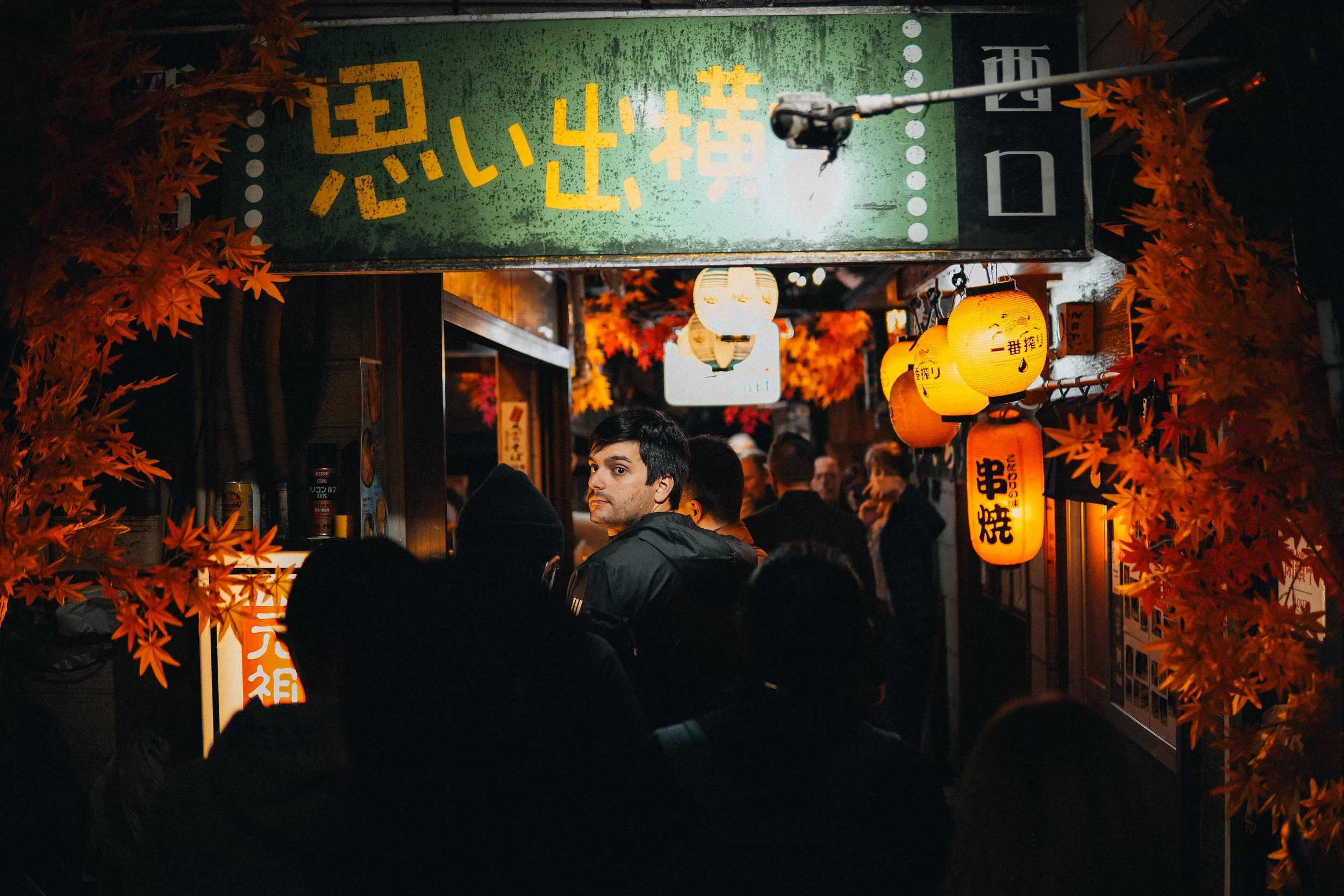 Night scene at a Japanese street food stall decorated with orange lanterns and red autumn leaves. A man with dark hair and a black jacket stands among a crowd of people.