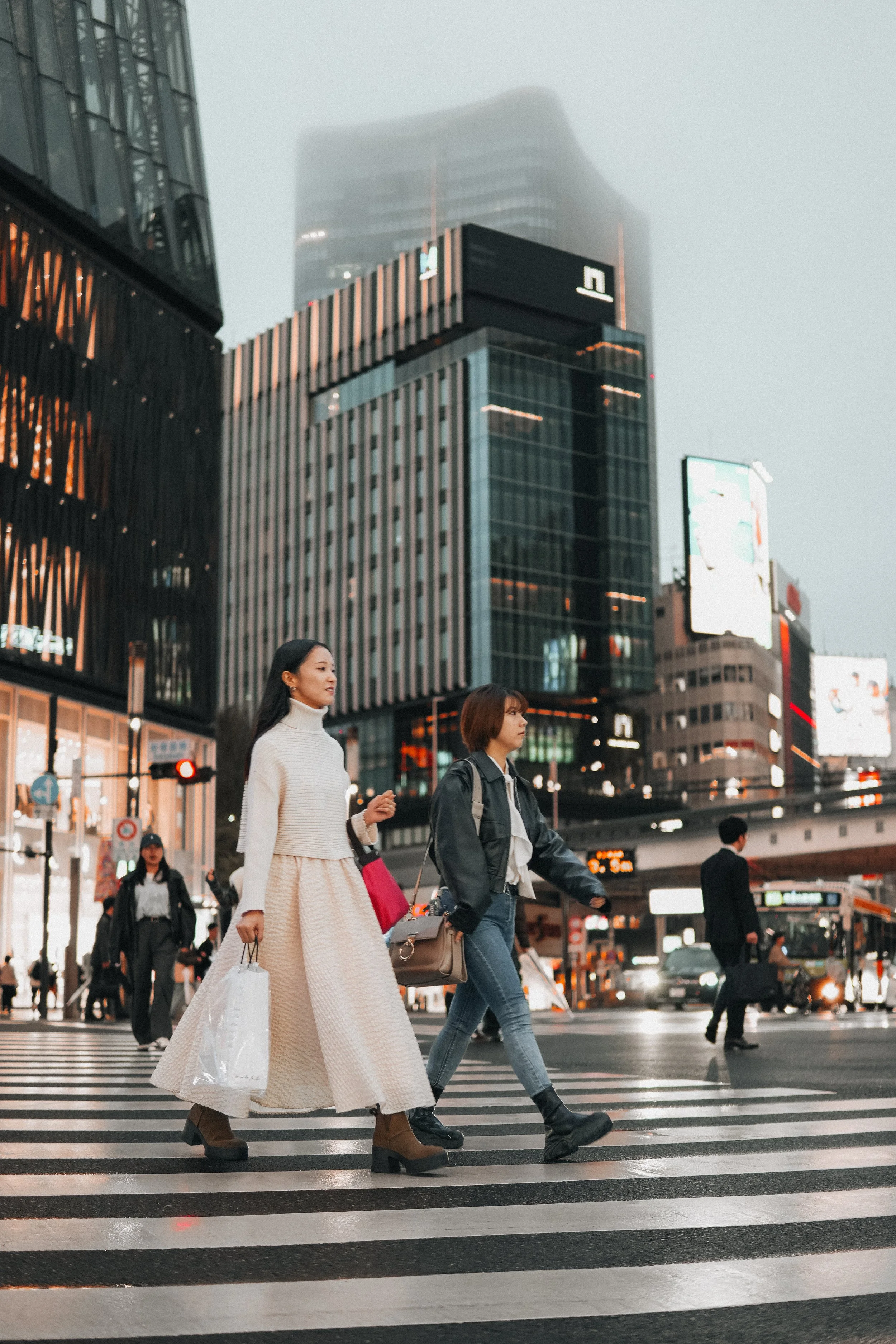 People crossing a busy city street at dusk with tall modern buildings in the background.