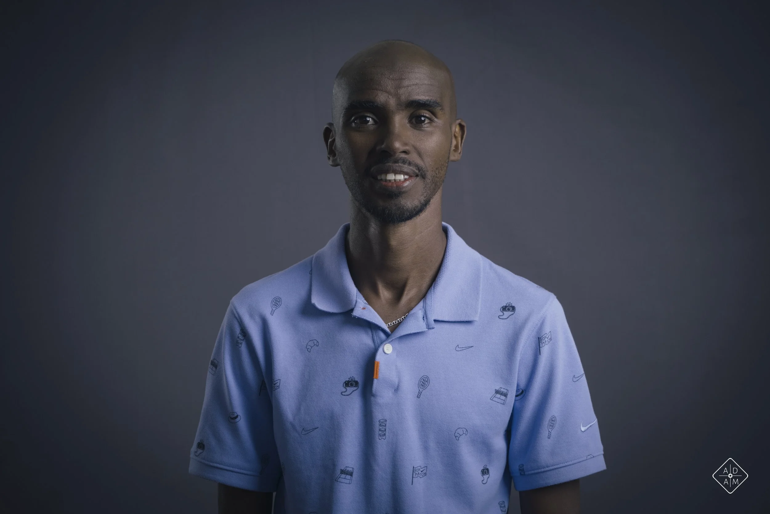 A young man with a shaved head wearing a light blue Nike polo shirt with small sports-themed icons against a dark background.