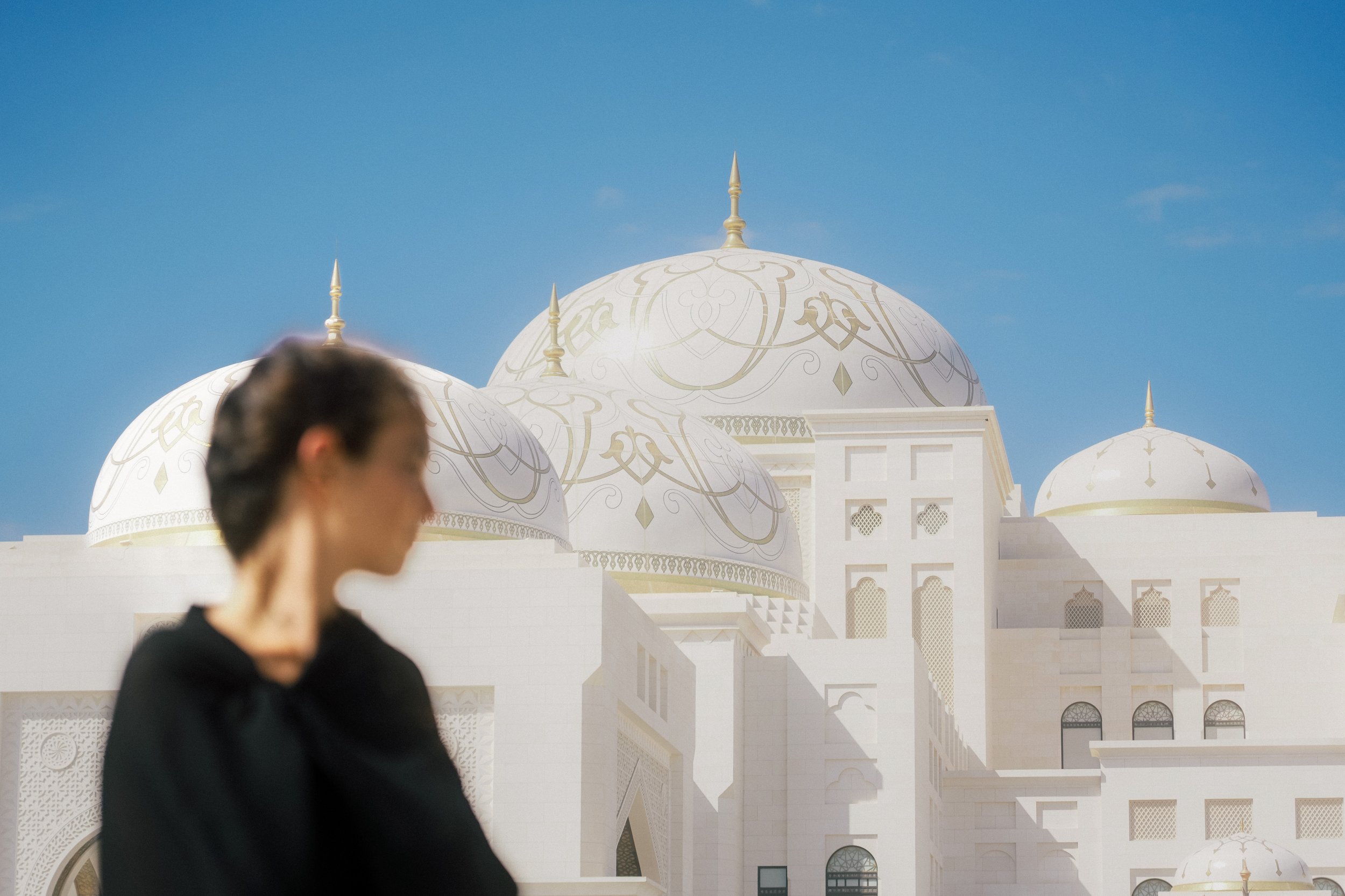 Blurred woman with short hair in black stands in front of a white building with domes and gold accents against a blue sky.