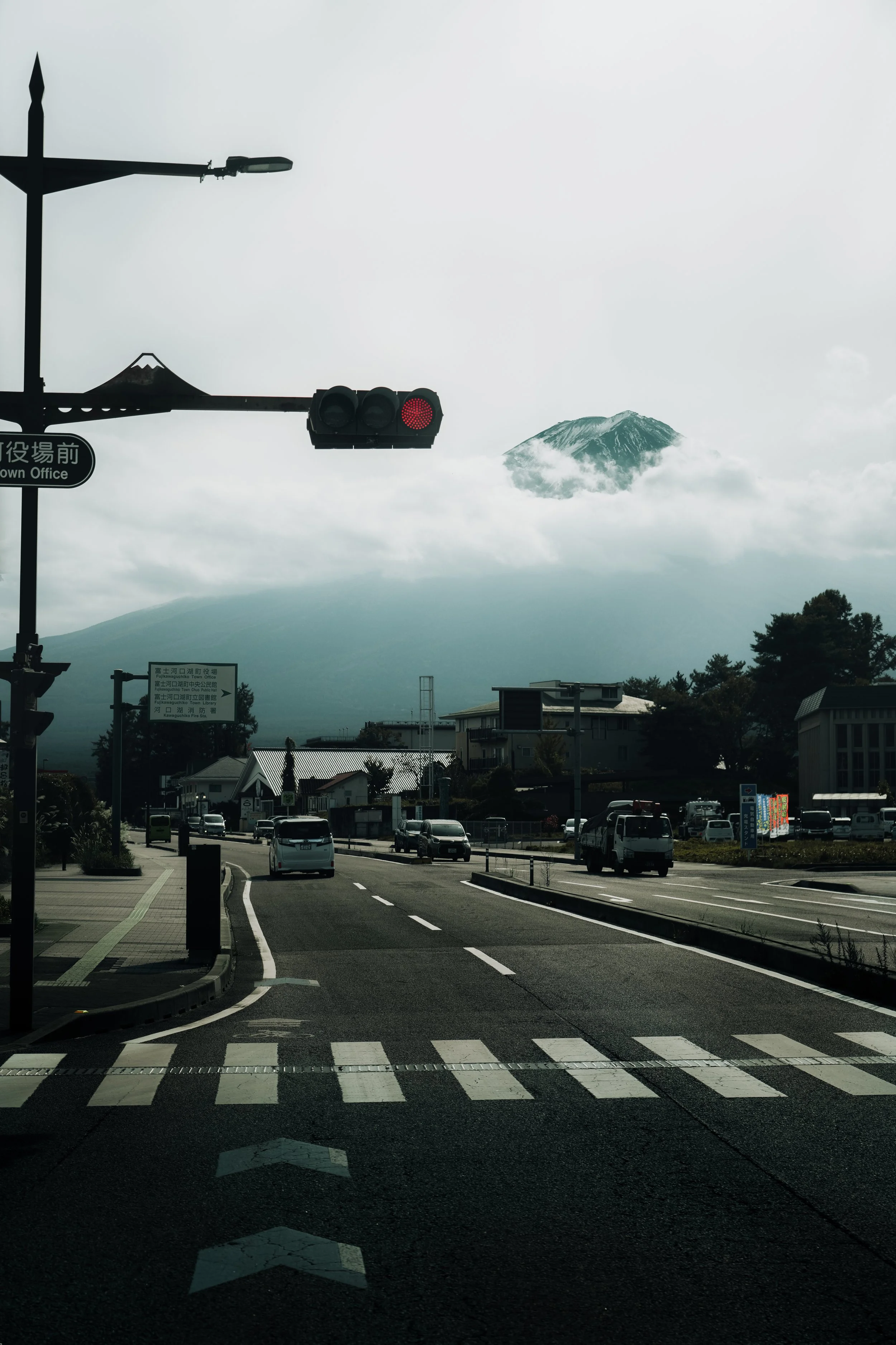 A city street with cars, traffic light, and signage, with Mount Fuji partially covered by clouds in the background.