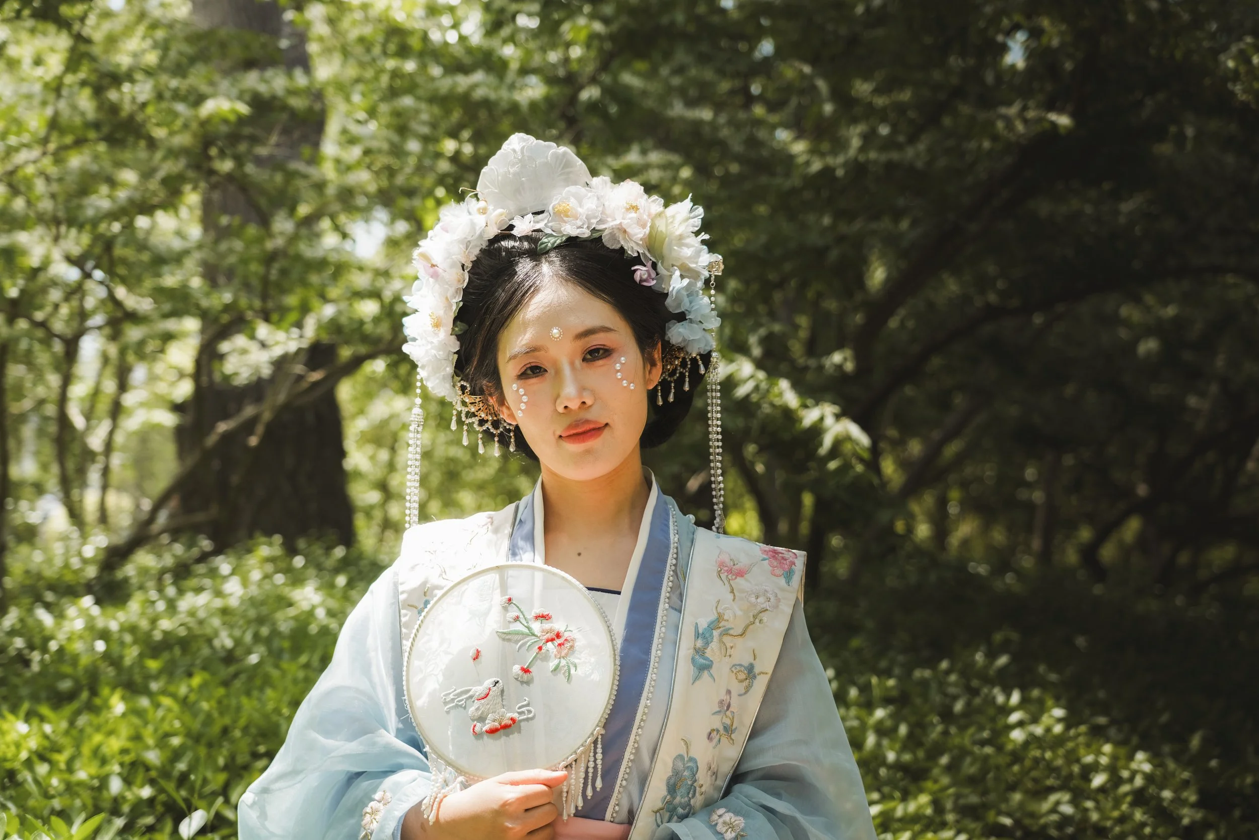 A woman dressed in traditional Asian attire, holding a decorative embroidered fan while standing in a lush green outdoor setting.