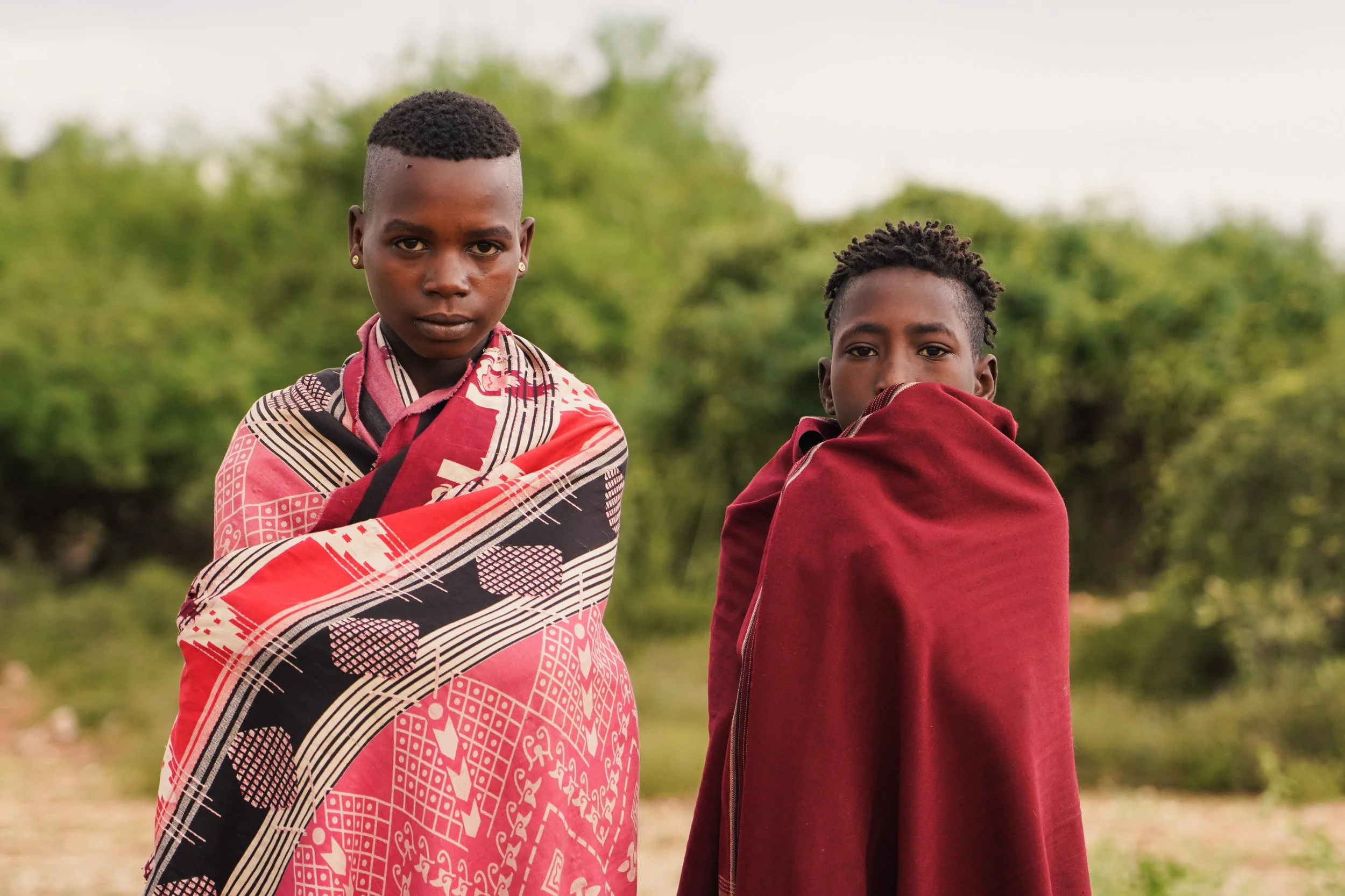 Two young children standing outdoors in traditional Maasai attire, wrapped in colorful shukas, with green trees in the background.