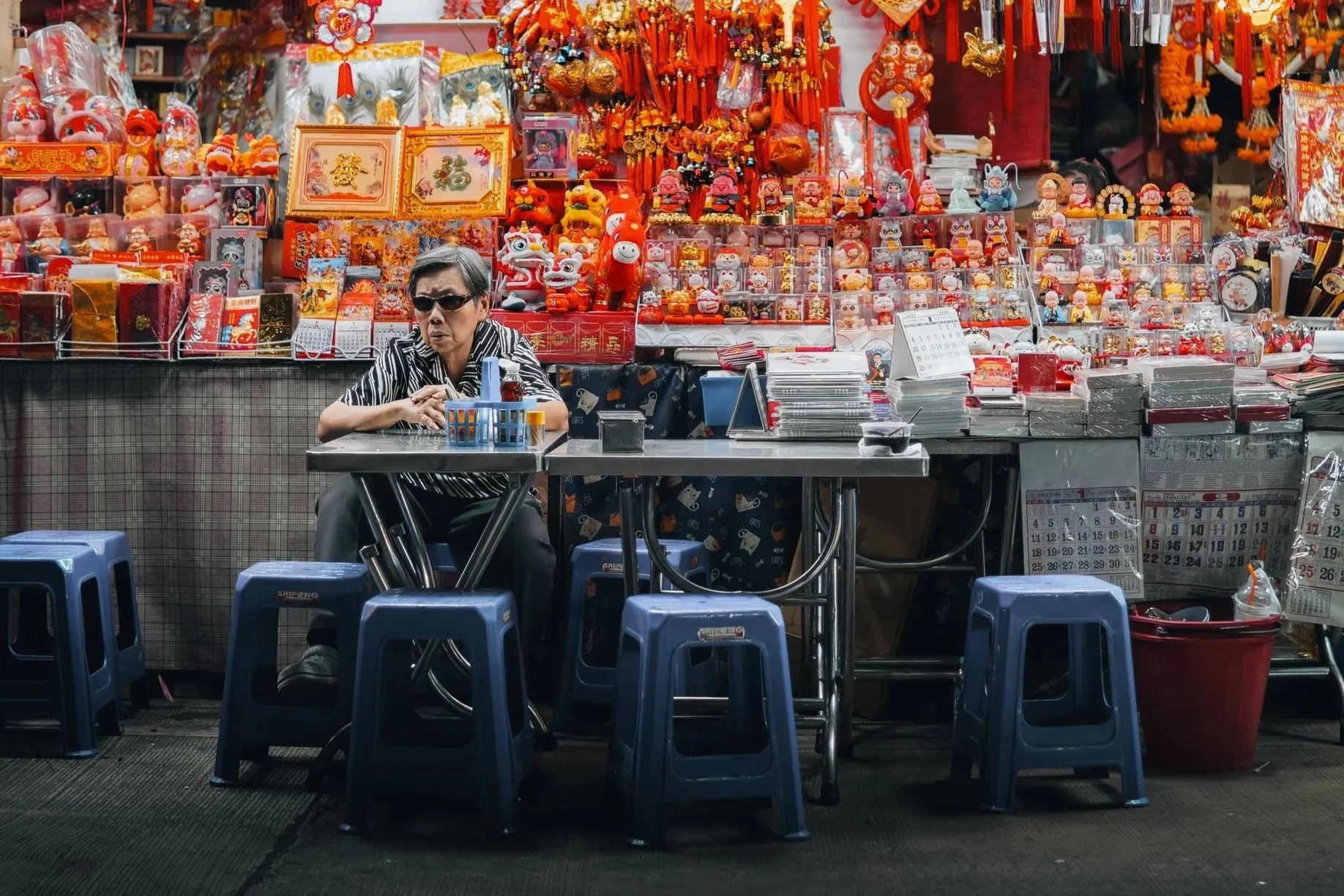 An elderly person sitting at a small table with multiple blue plastic stools in front of a colorful market stall decorated with various Chinese New Year decorations, figurines, and red ornaments. The person is wearing sunglasses and a striped shirt.