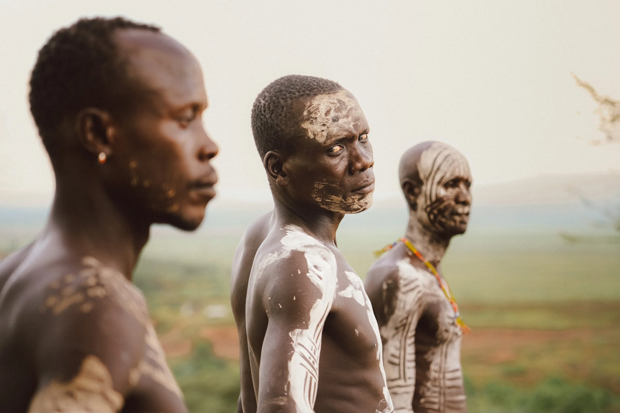 Three indigenous men with traditional face and body paint standing outdoors in an open landscape.