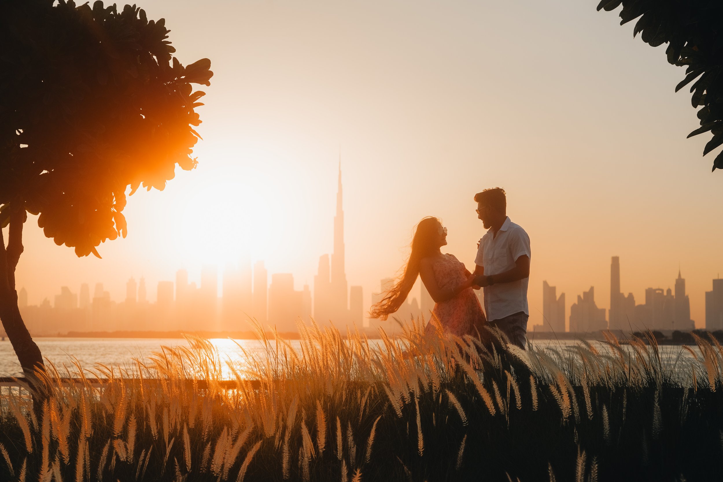A couple holding hands and looking at each other near a body of water with a city skyline and the Burj Khalifa in the background during sunset.