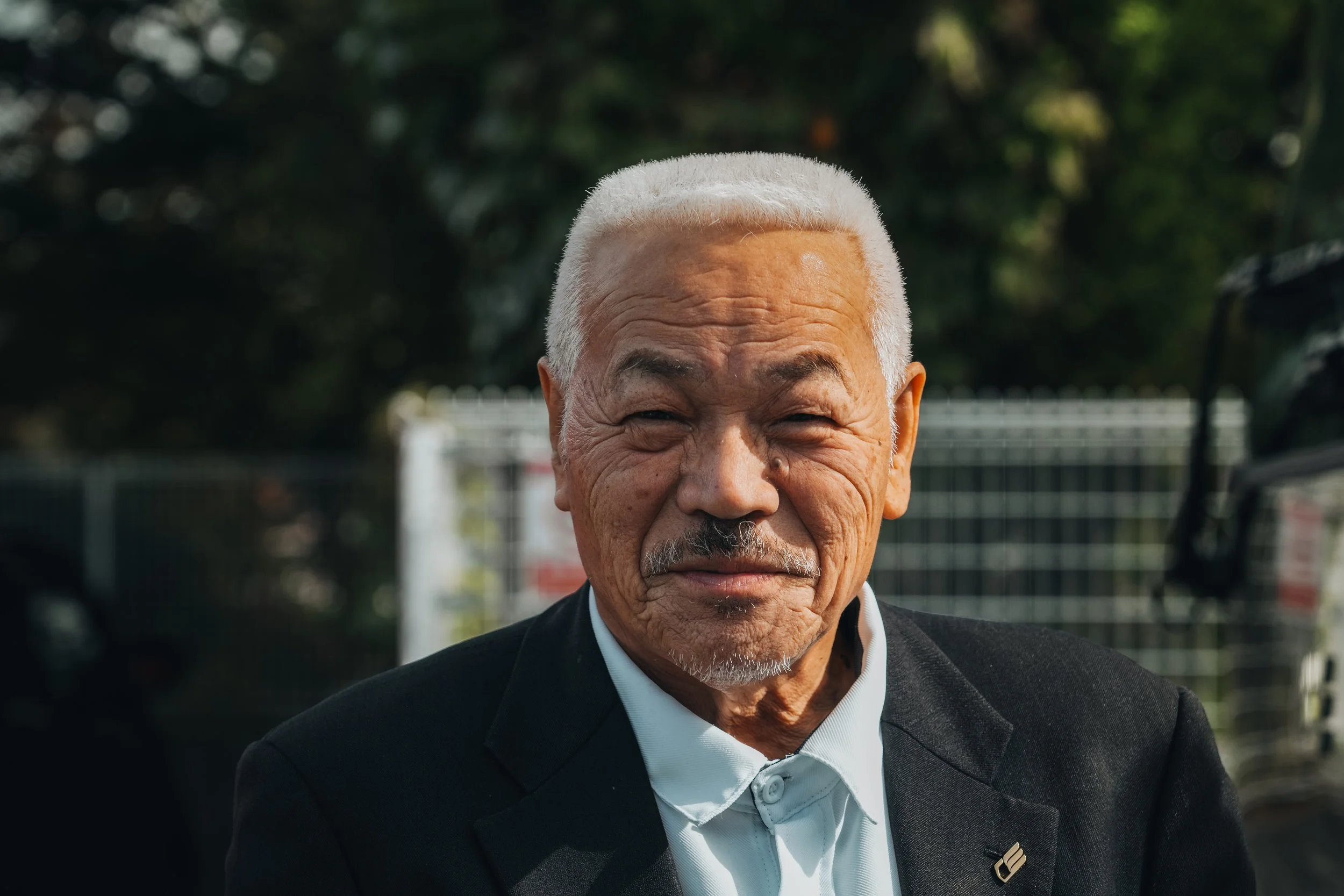 An elderly man with white hair and a mustache, wearing a black blazer and white shirt, smiling outdoors with blurred trees and a fence in the background.