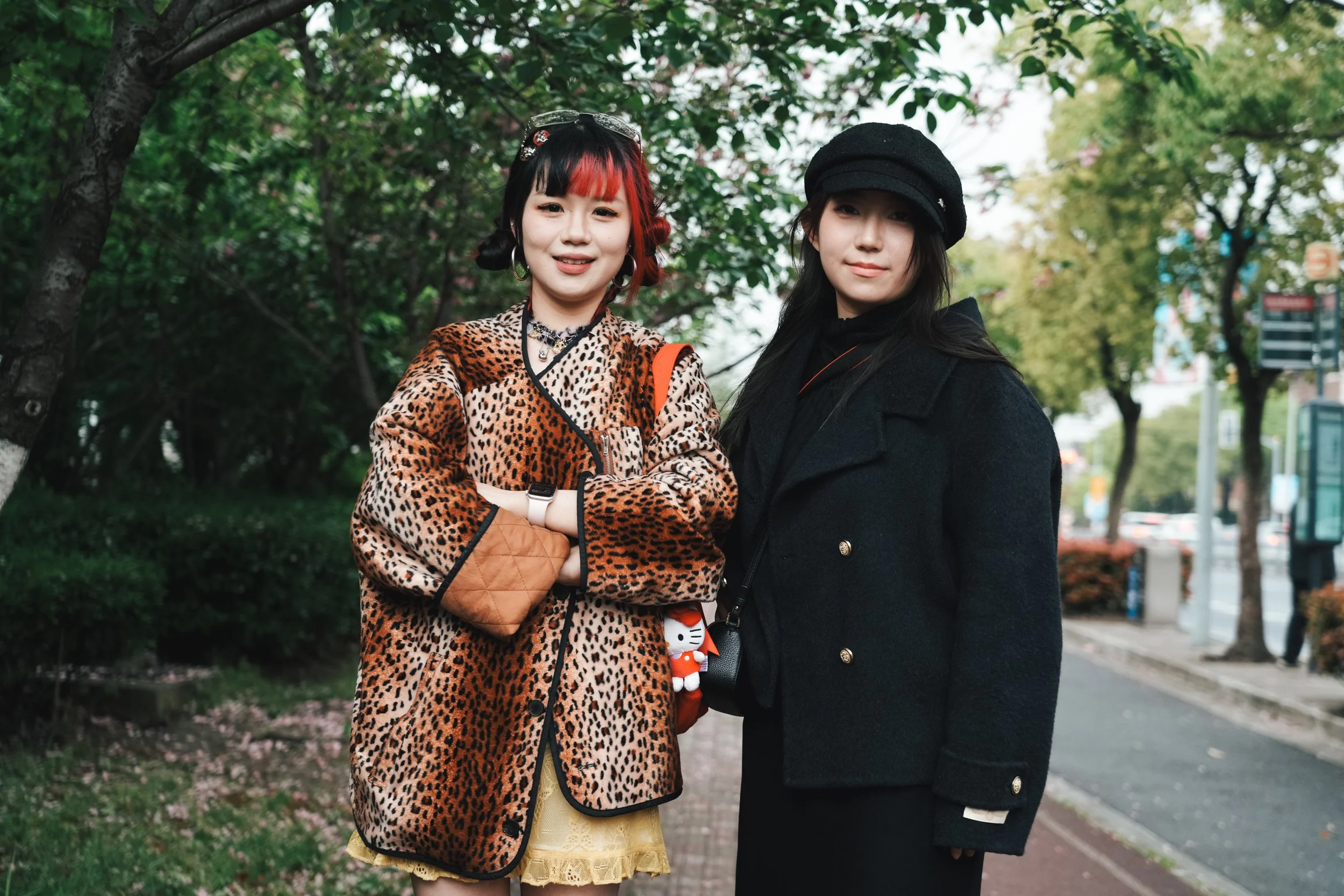 Two young women standing together outdoors on a city sidewalk with trees and buildings in the background. One woman wears a leopard print jacket, a yellow dress, and has red and black hair, while the other wears a black coat and hat with long dark ha