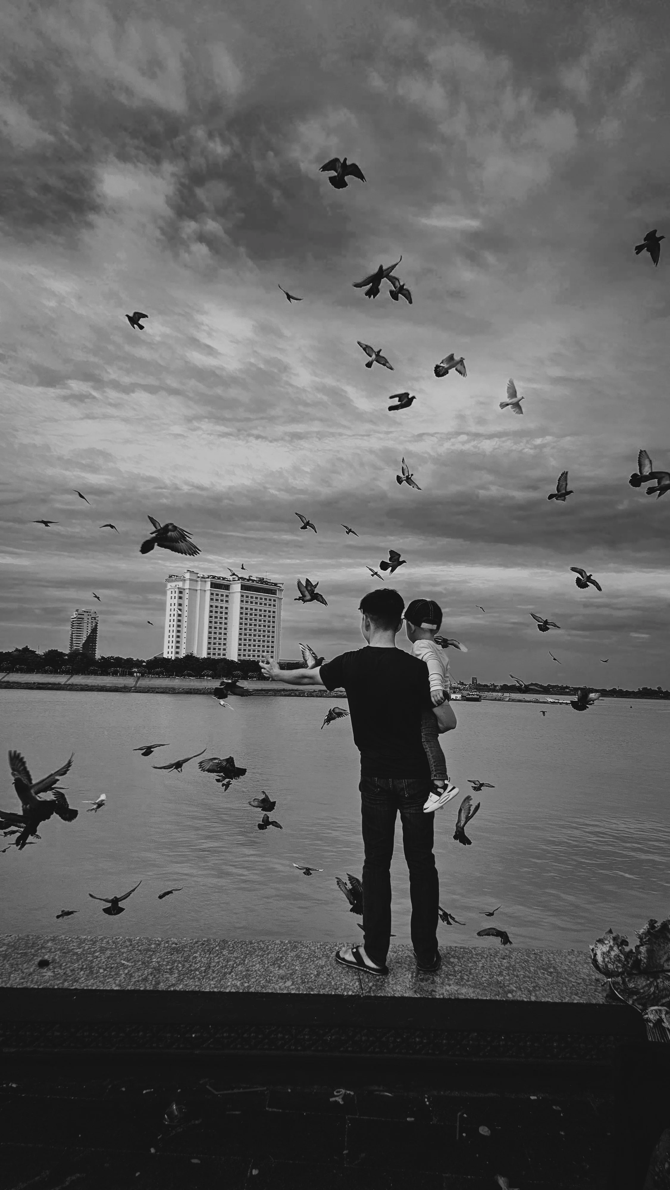 A person holding a child by a body of water. Their back is to the camera. Birds are flying in the sky around them, with buildings in the background. The image is in black and white.