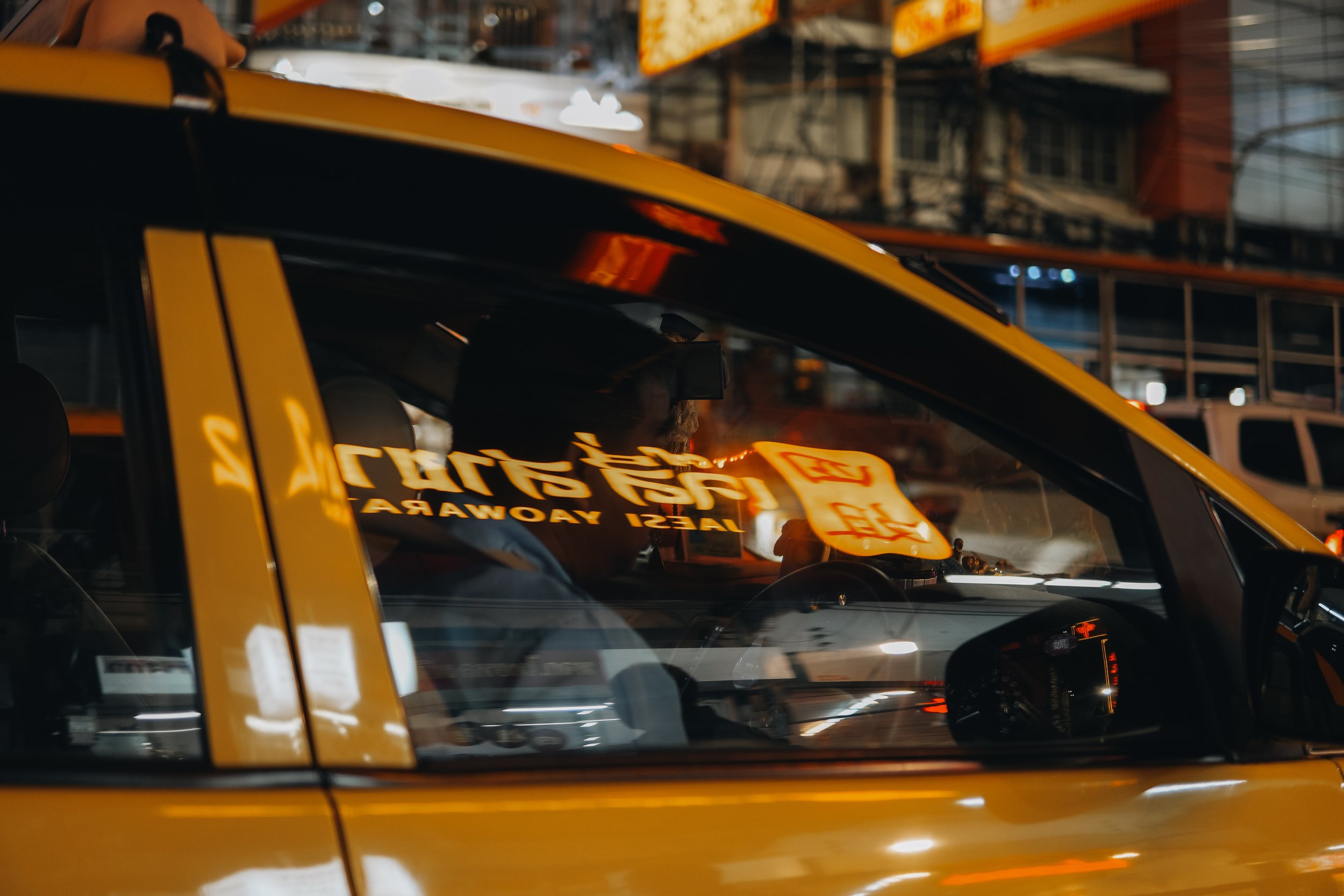 A taxi driver sitting inside a yellow taxi vehicle at night, with reflections of neon signs in the window, in an urban area.