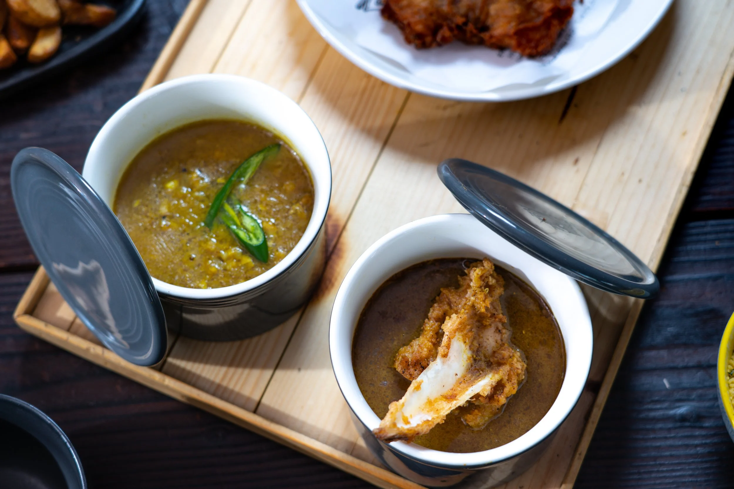 Two bowls of Indian curry with lids open, one containing yellow lentil curry topped with green chili, the other with a chicken curry topped with a piece of chicken, placed on a wooden tray.