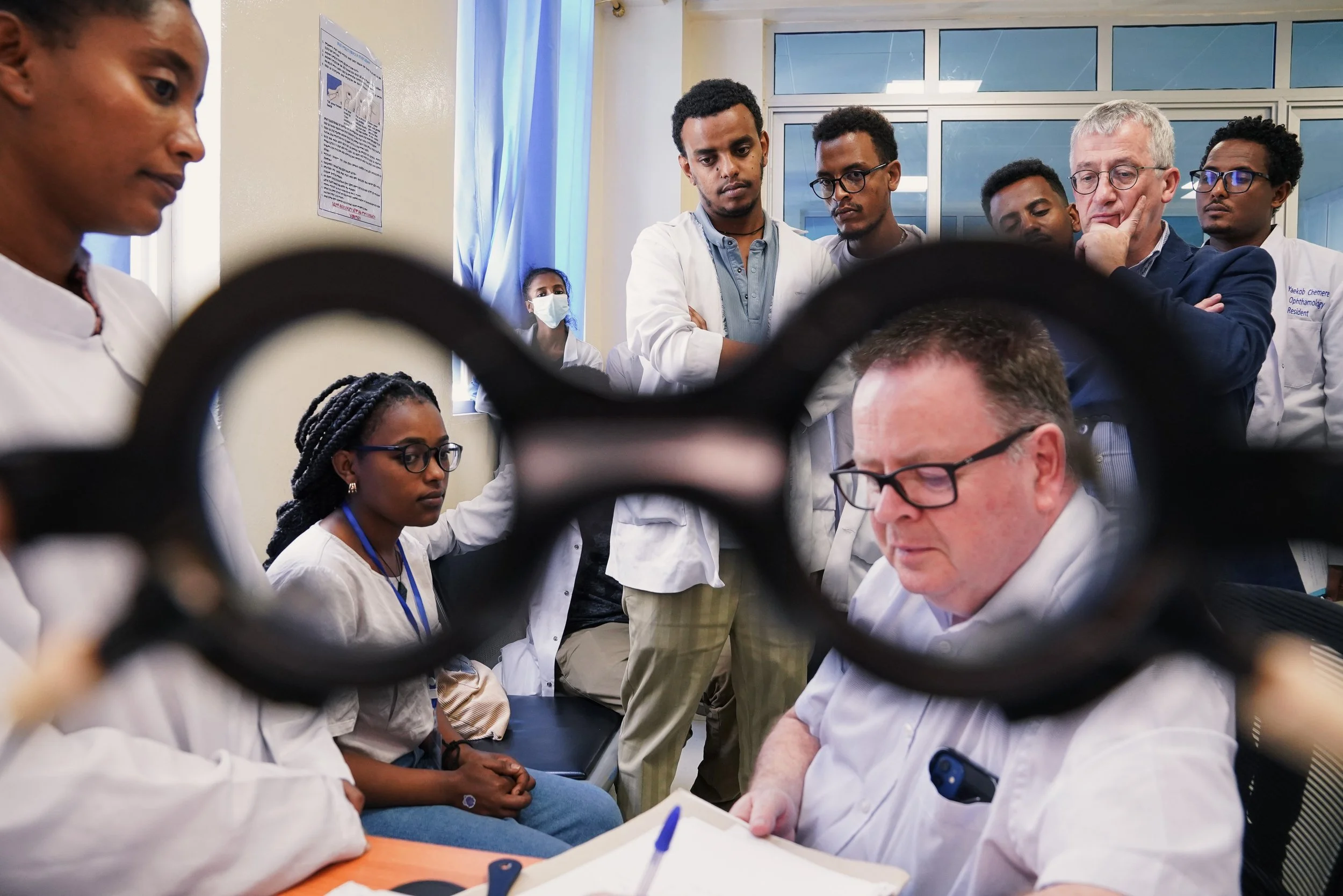 Medical professionals and students gathered around a doctor in a hospital or clinic, with a pair of glasses in the foreground.