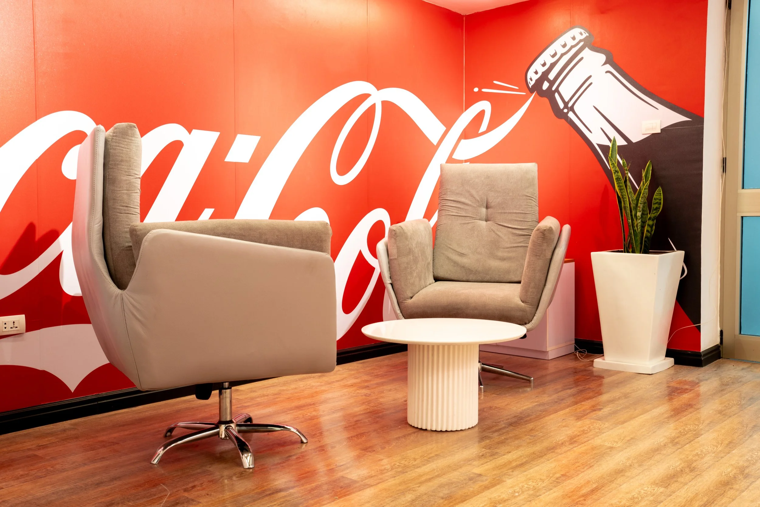 Interior lounge area with Coca-Cola mural, two beige chairs, a white side table, and a potted plant on a wood floor.