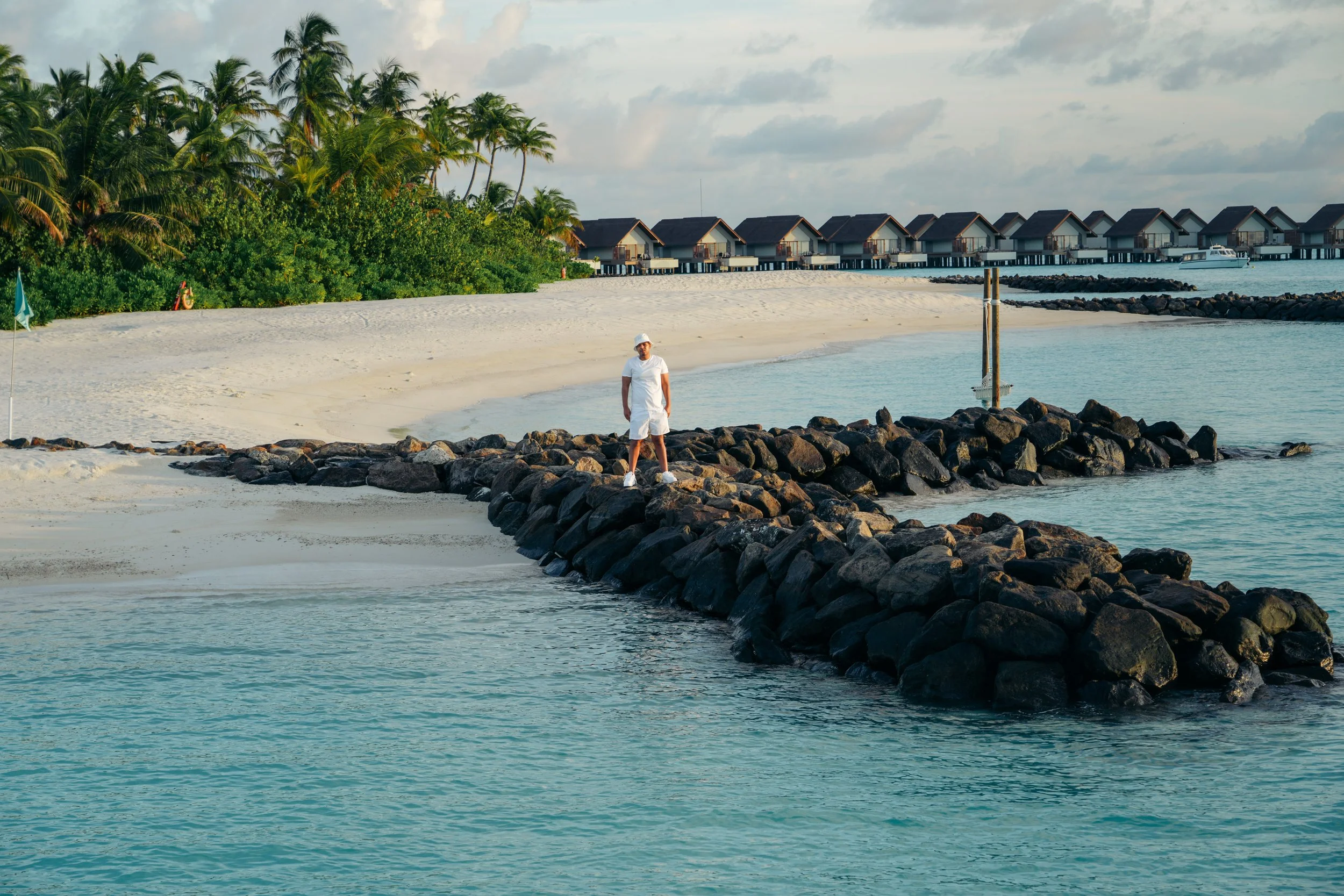 A man in white clothing and a hat standing on a rocky jetty on a tropical beach with waves, white sand, palm trees, and overwater bungalows in the background.