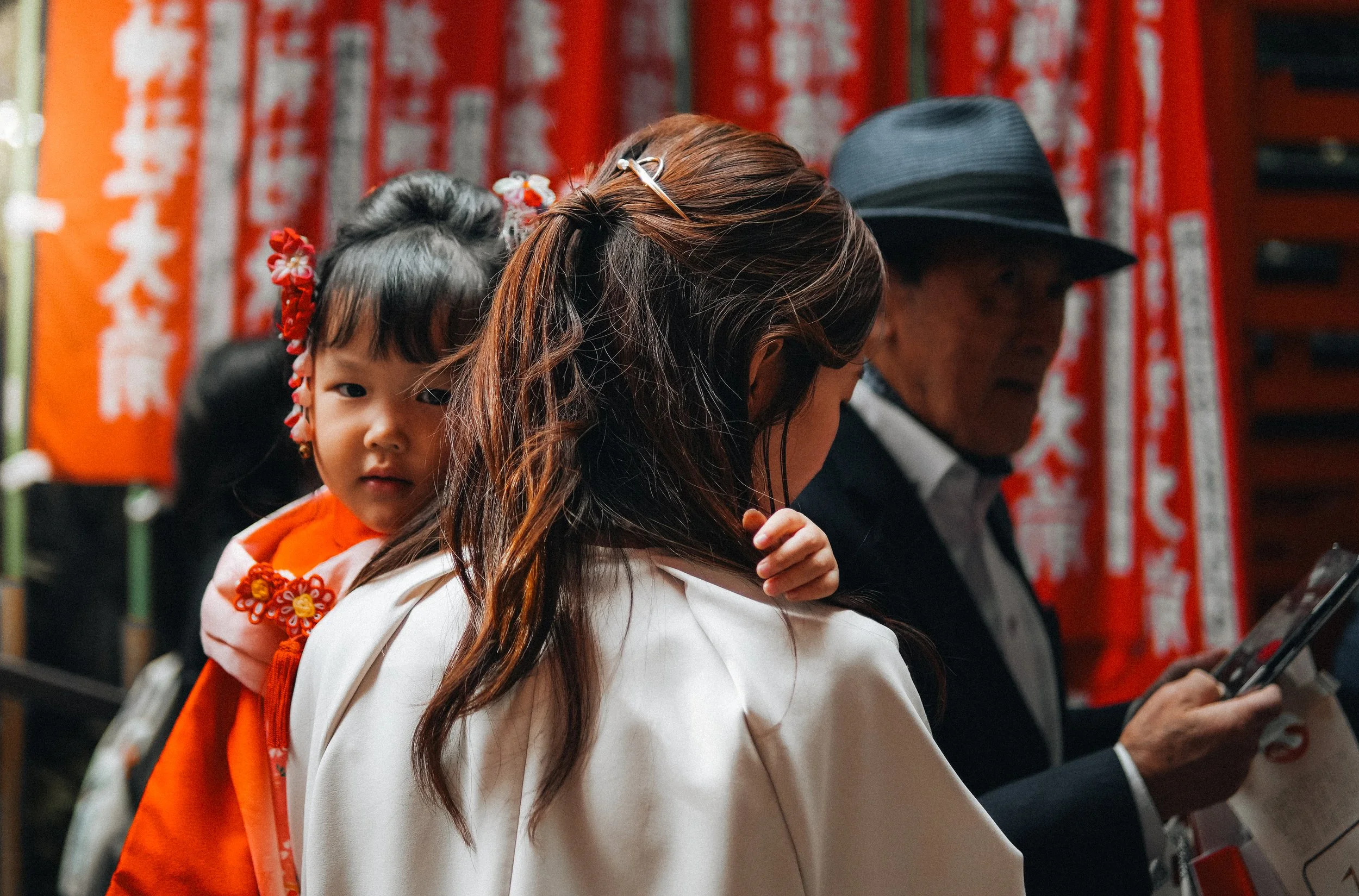 A young girl dressed in traditional Japanese attire with colorful decorations in her hair, looks directly at the camera while being carried on a woman's back. The woman has long wavy hair and is wearing a white garment. An elderly man in a hat and su