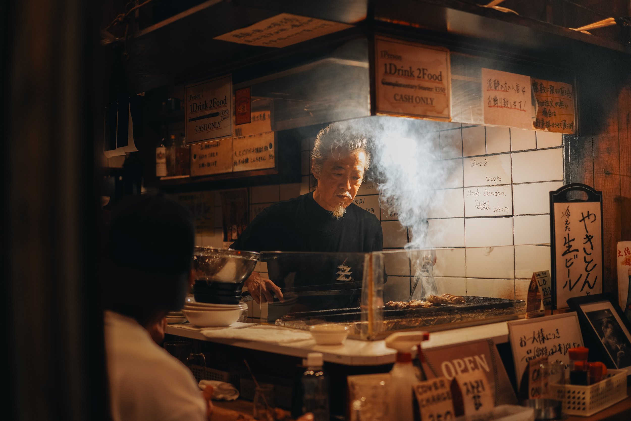 An older man with gray hair and a beard cooking at a food stall, with smoke rising from the grill, inside a Japanese eatery with handwritten signs in Japanese and English.