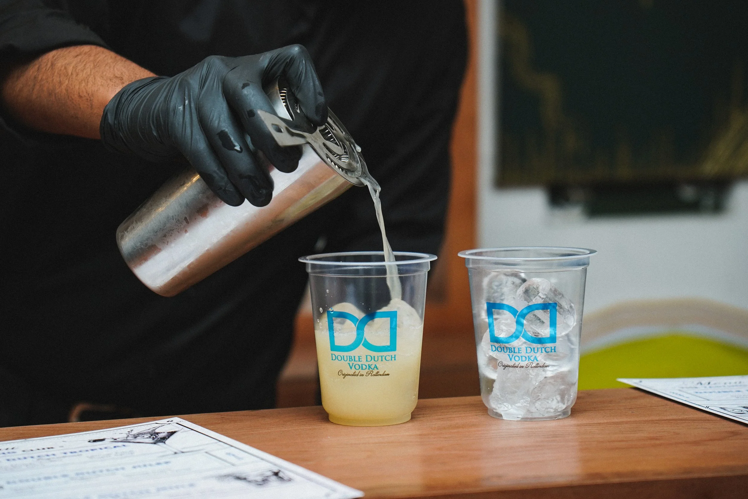 Person wearing black gloves pouring a yellow-colored liquid from a can into a clear plastic cup with the Double Dutch Vodka logo, next to another cup with ice, on a bar counter.