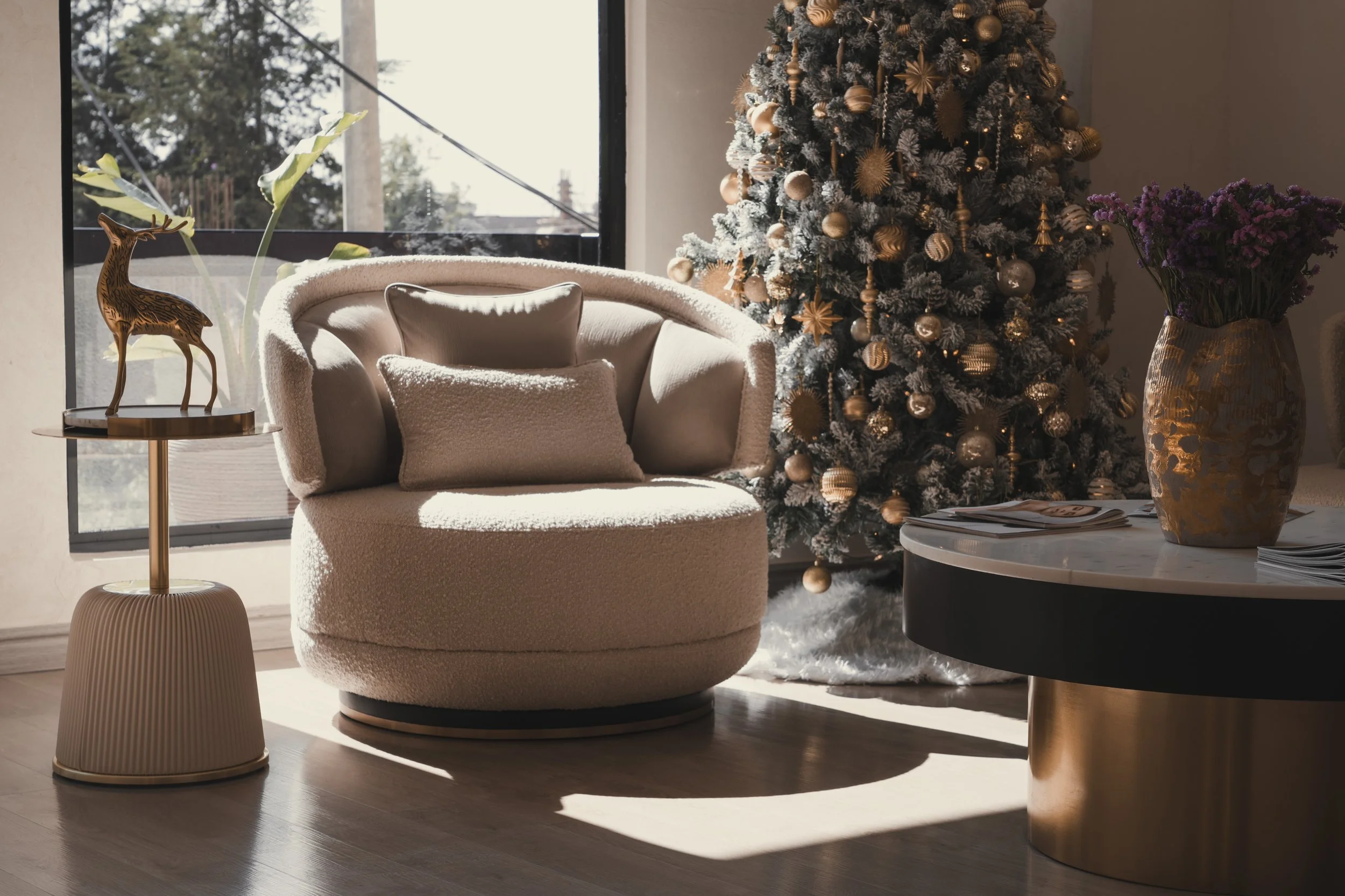 Living room decorated for Christmas with a Christmas tree, a cream armchair with cushions, a side table with a decorative reindeer and plants, and a round dining table with a vase of purple flowers.