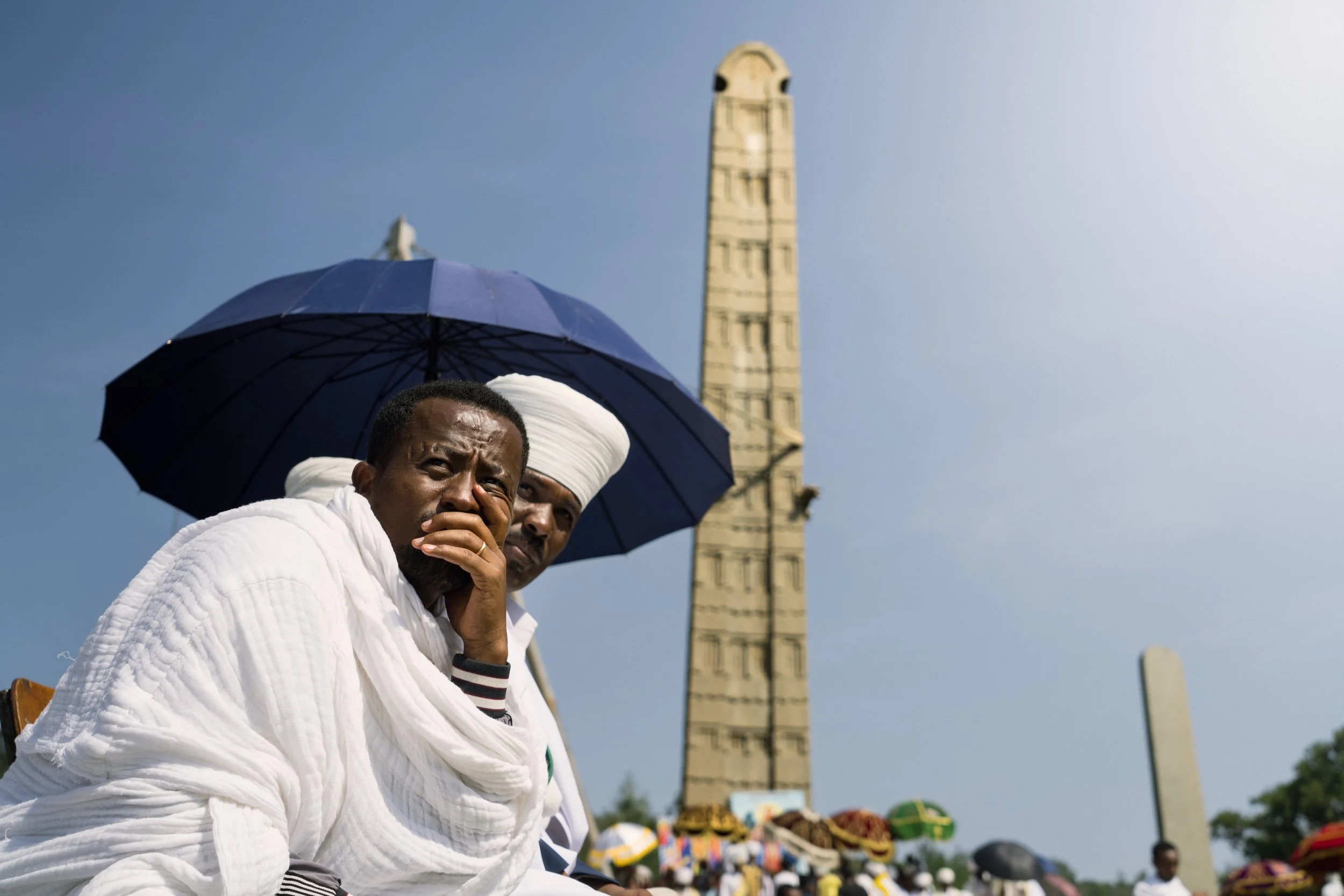 Two men in white traditional clothing sitting outdoors under a dark umbrella, with a tall monument and a gathering of people with umbrellas in the background.