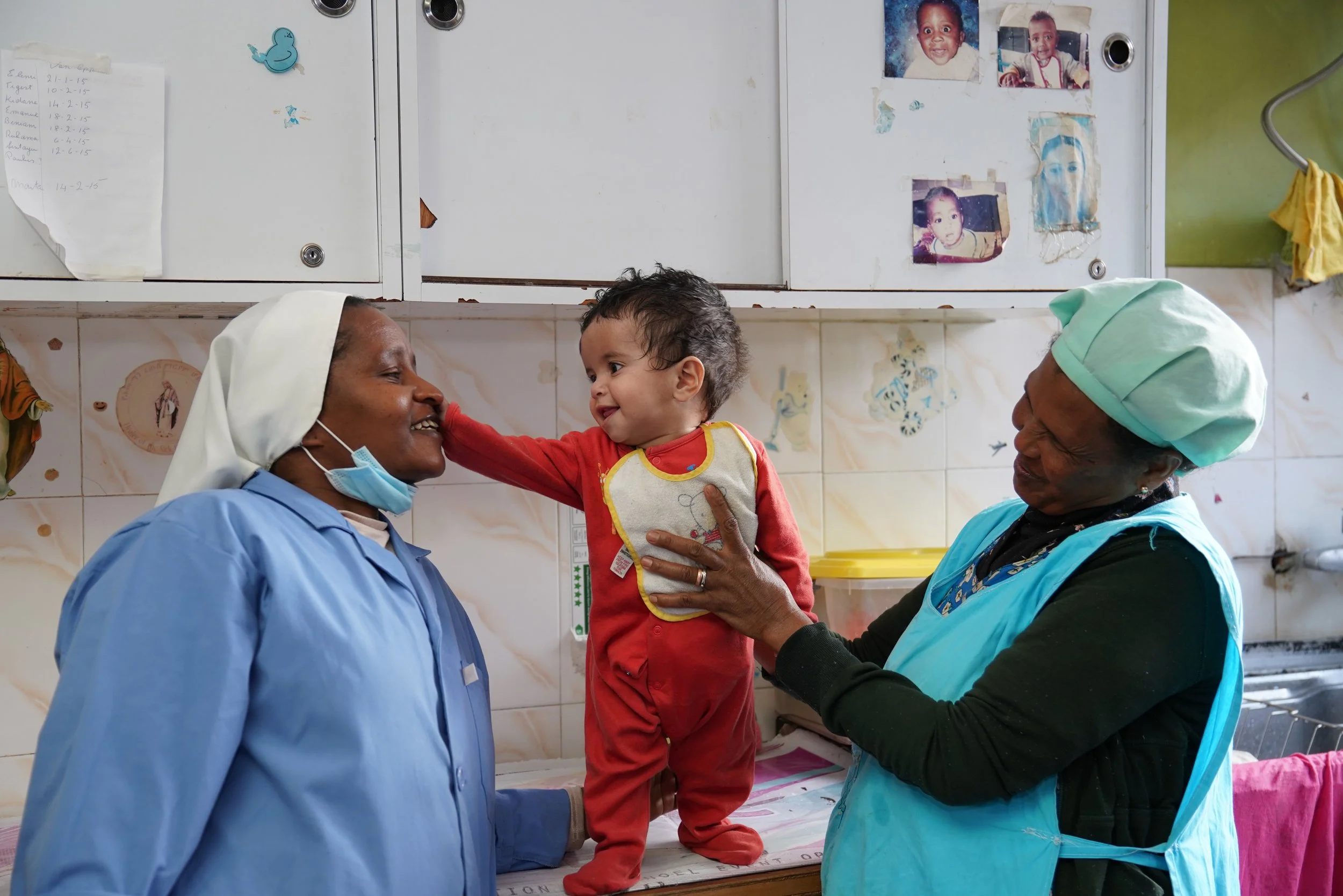 A young child in red outfit and bib is being held up by a woman in medical scrubs, with another nurse beside her. The child is touching the woman’s face, and they are all smiling. The setting appears to be a clinic or hospital room with photos and no