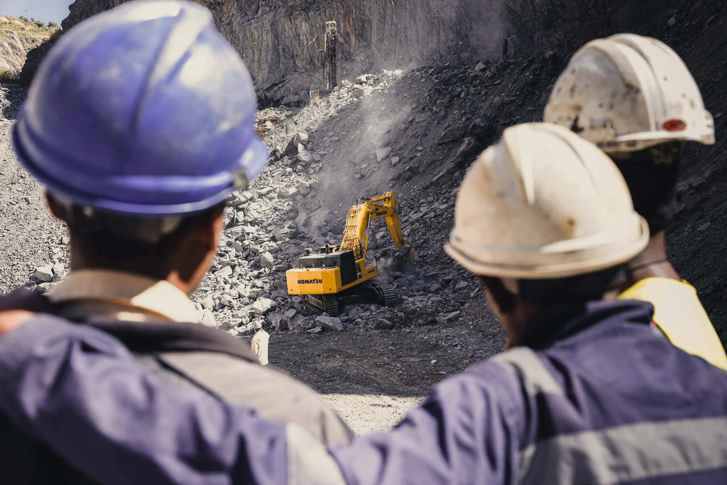 Three construction workers wearing helmets observe a yellow excavator working on a rocky slope in a mining or construction site.