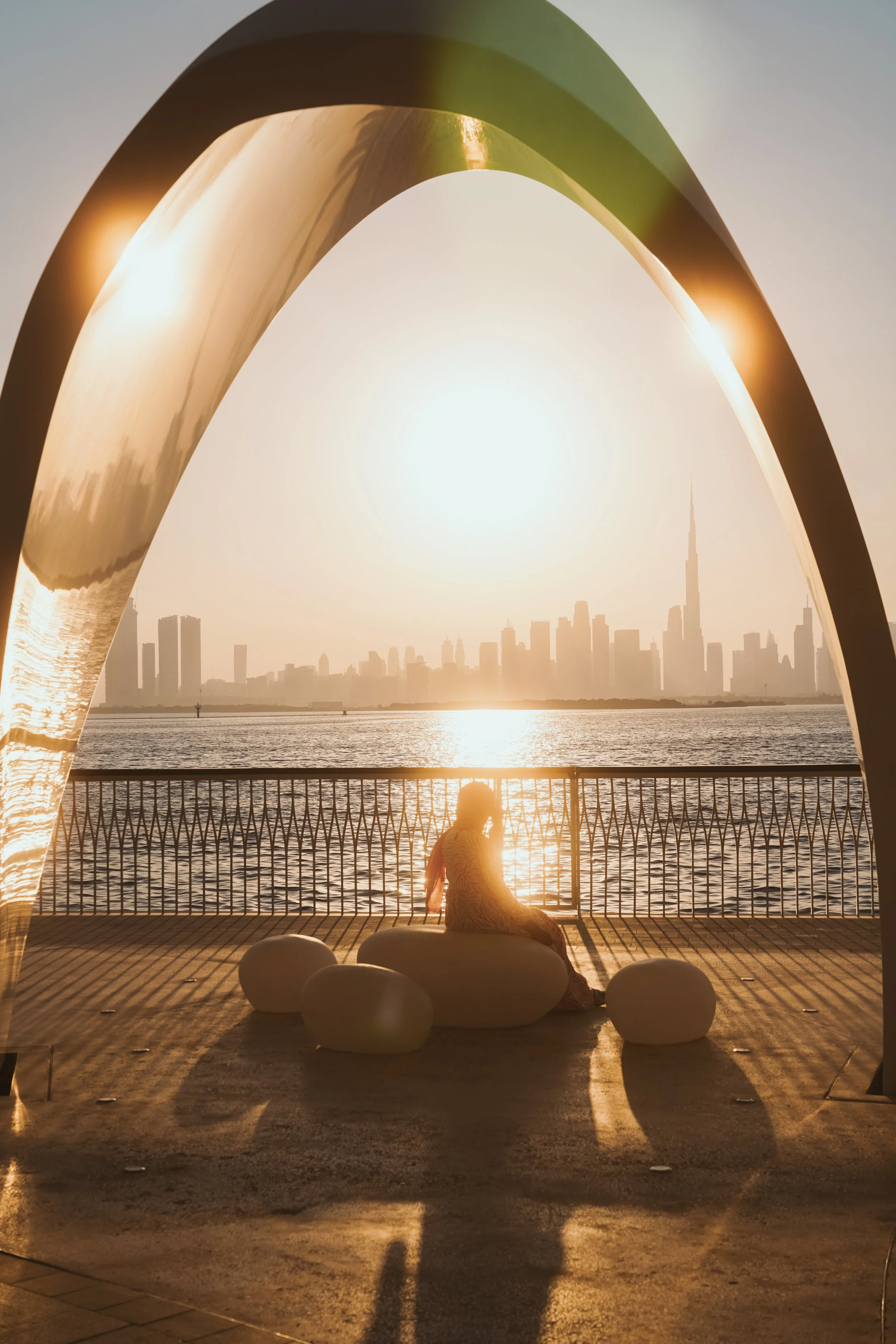 A woman sitting on a bench by the water during sunset, viewed through a modern metal arch with a city skyline in the background.
