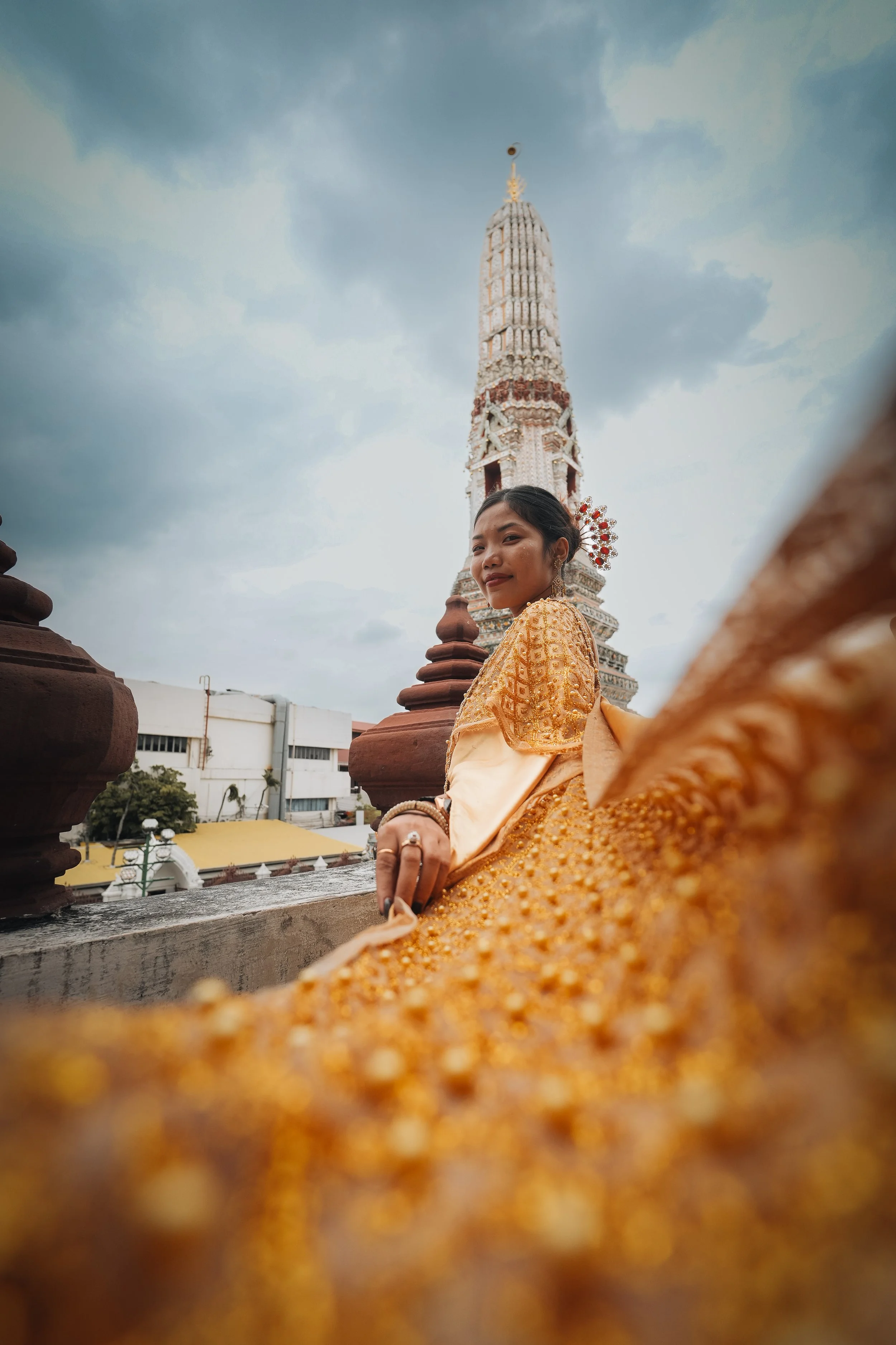 A woman dressed in traditional gold attire sitting on a temple rooftop with a tall ornate spire in the background under a cloudy sky.