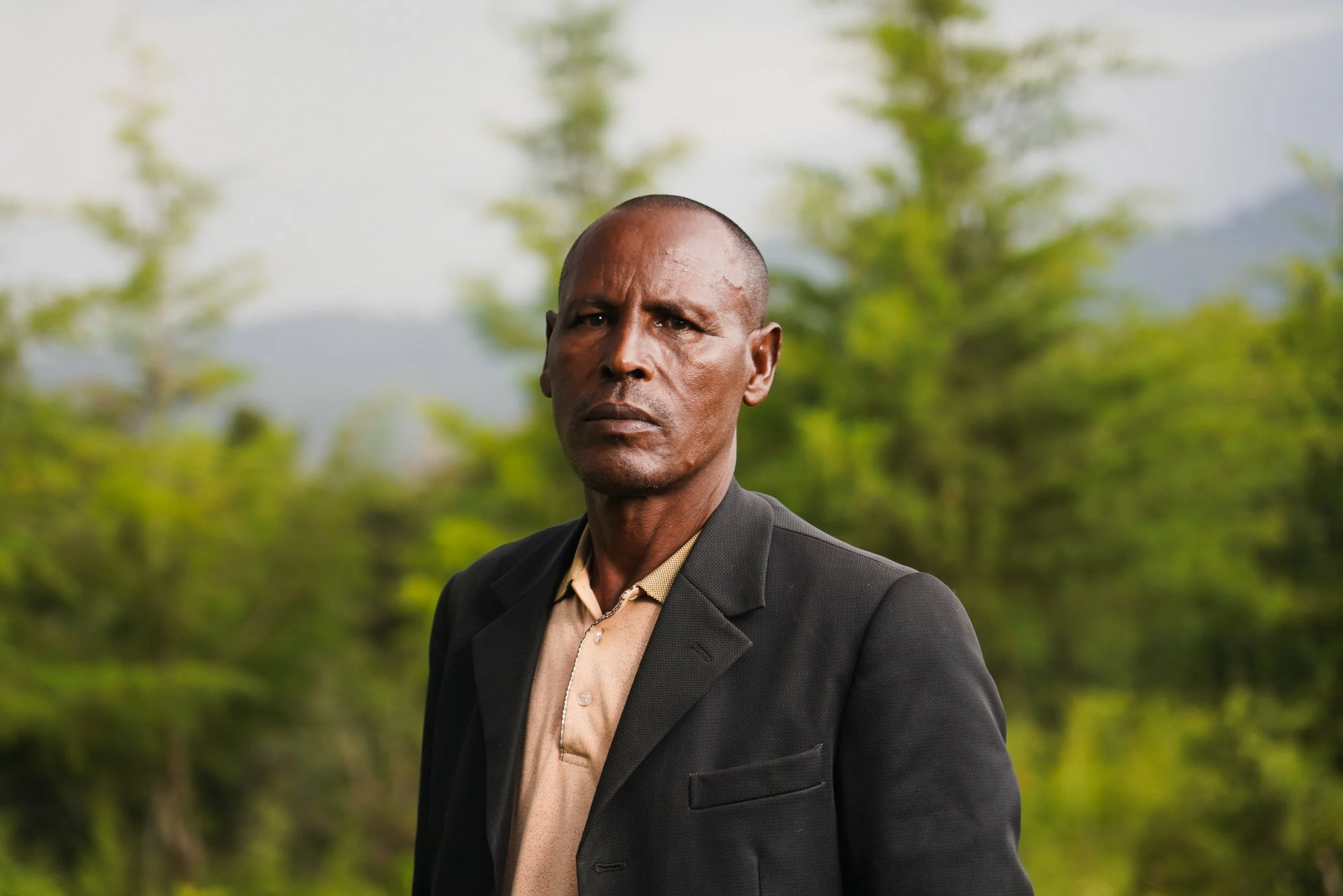 A stern-looking man with a serious expression, dressed in a black blazer and light-colored shirt, standing outdoors with blurred green trees and mountains in the background.