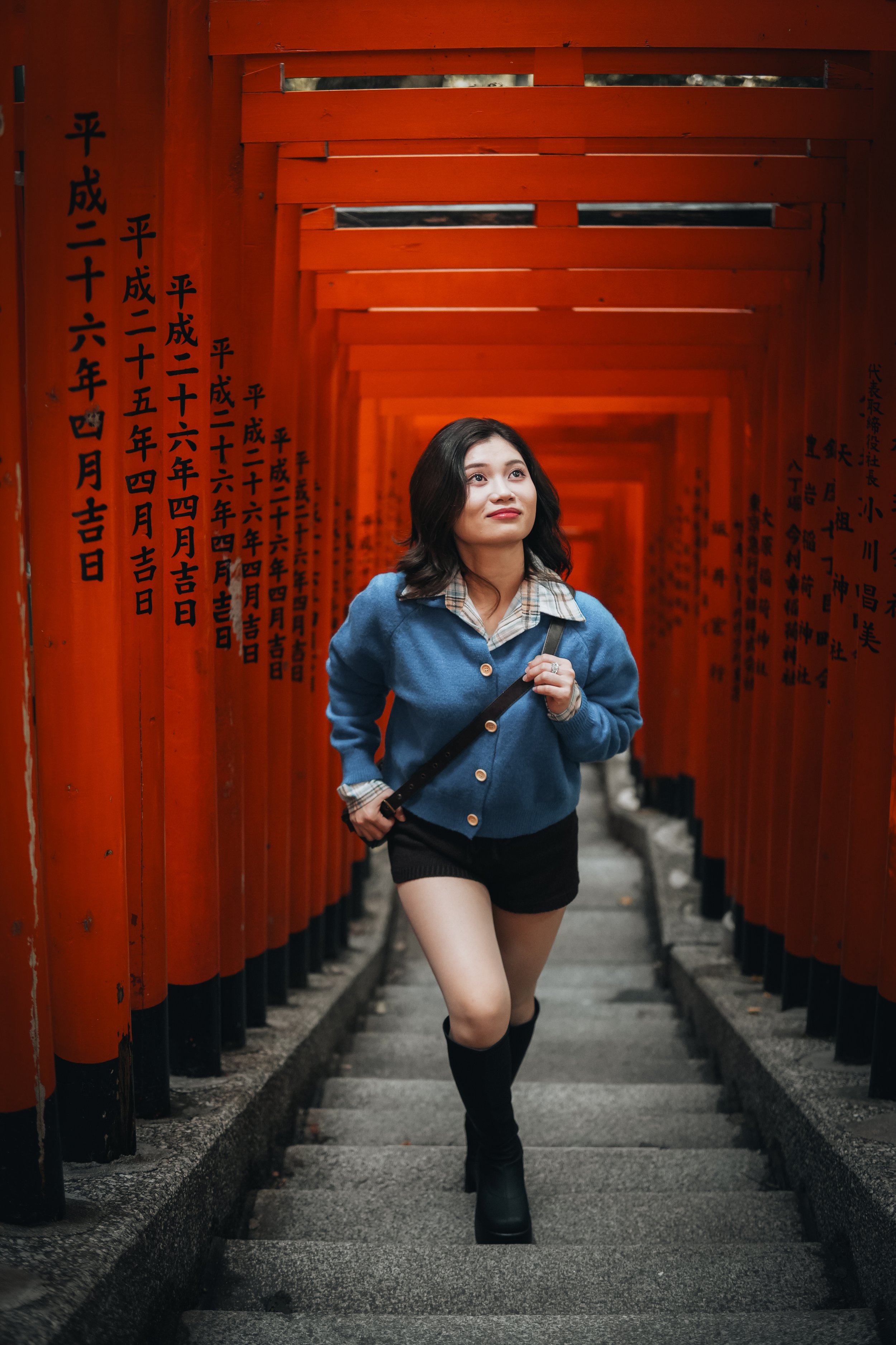A young woman walking through a corridor of red torii gates at Fushimi Inari Shrine in Japan, with black Japanese writing on the gates and wearing a blue sweater, plaid shirt, black shorts, and knee-high boots.