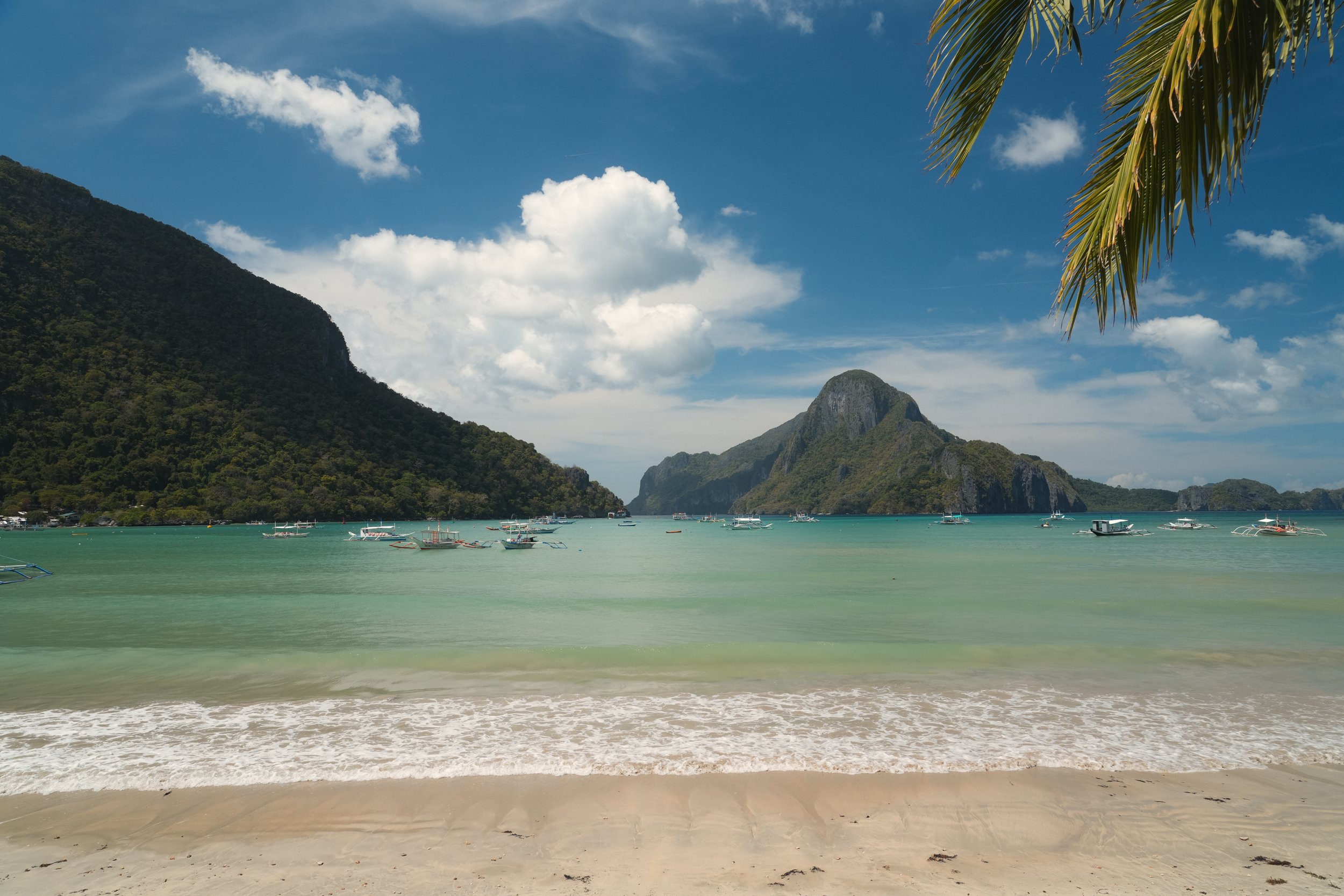 Tropical beach with white sand, calm turquoise water, boats floating on water, lush green hills in the background, blue sky with scattered clouds, and a palm tree branch in the upper right corner.