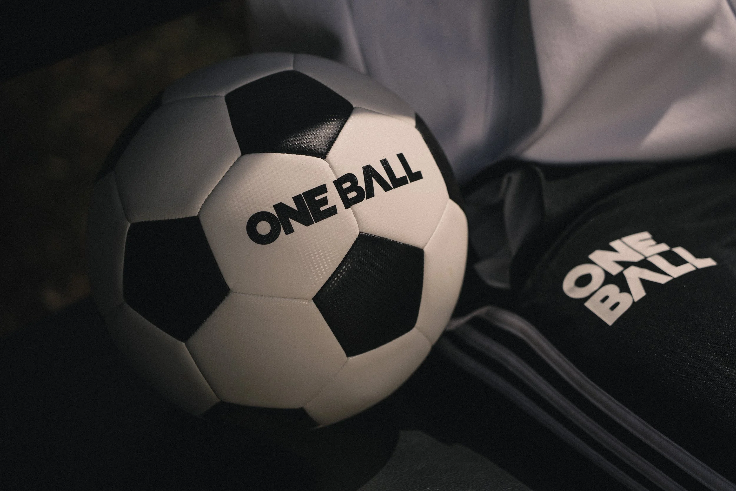 Close-up of a black and white soccer ball with the words 'ONE BALL' printed on it, resting against an open black bag with the same text in white.