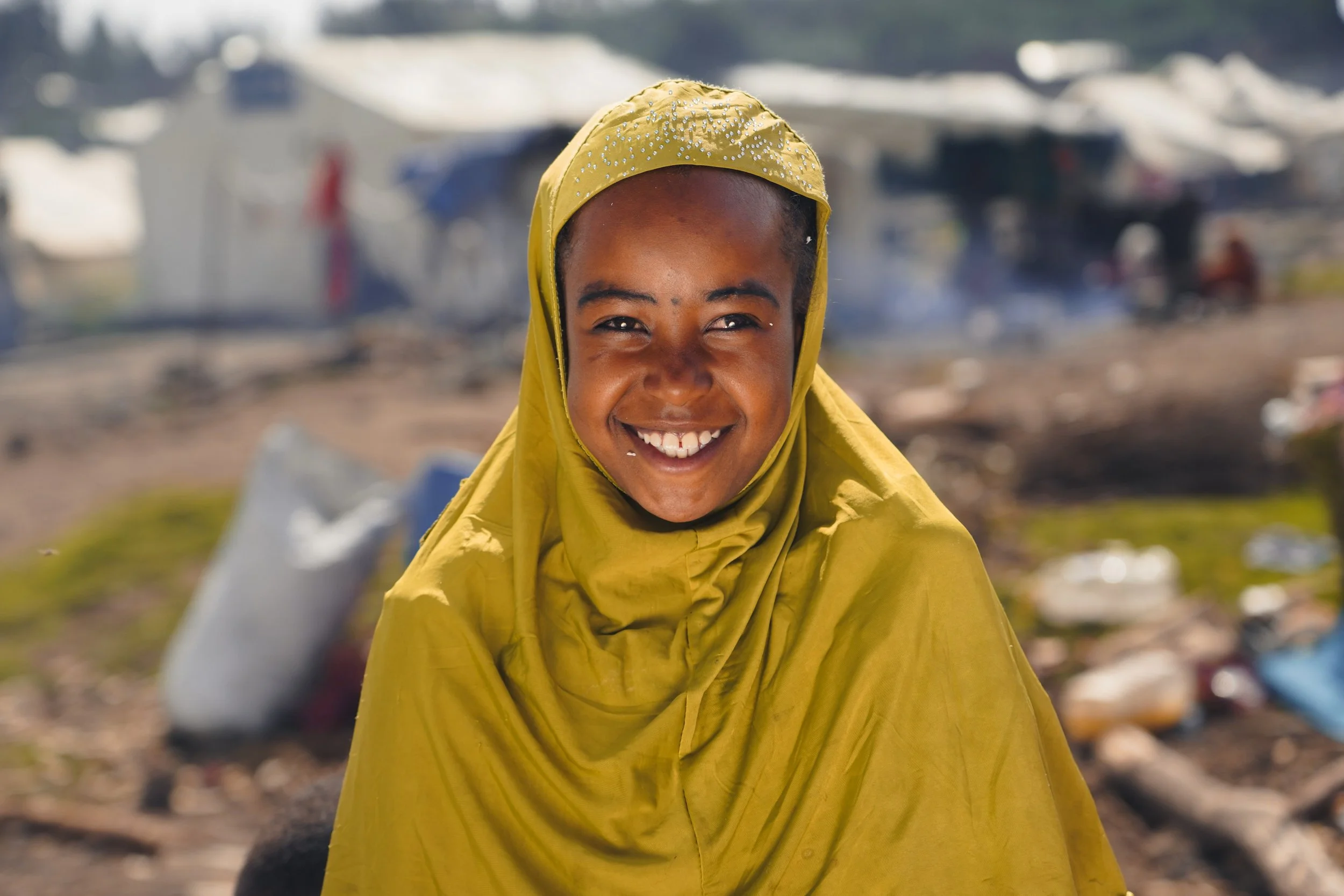 A smiling woman with dark skin, wearing a yellow headscarf and yellow clothing, outdoors with blurred tents and debris in the background.