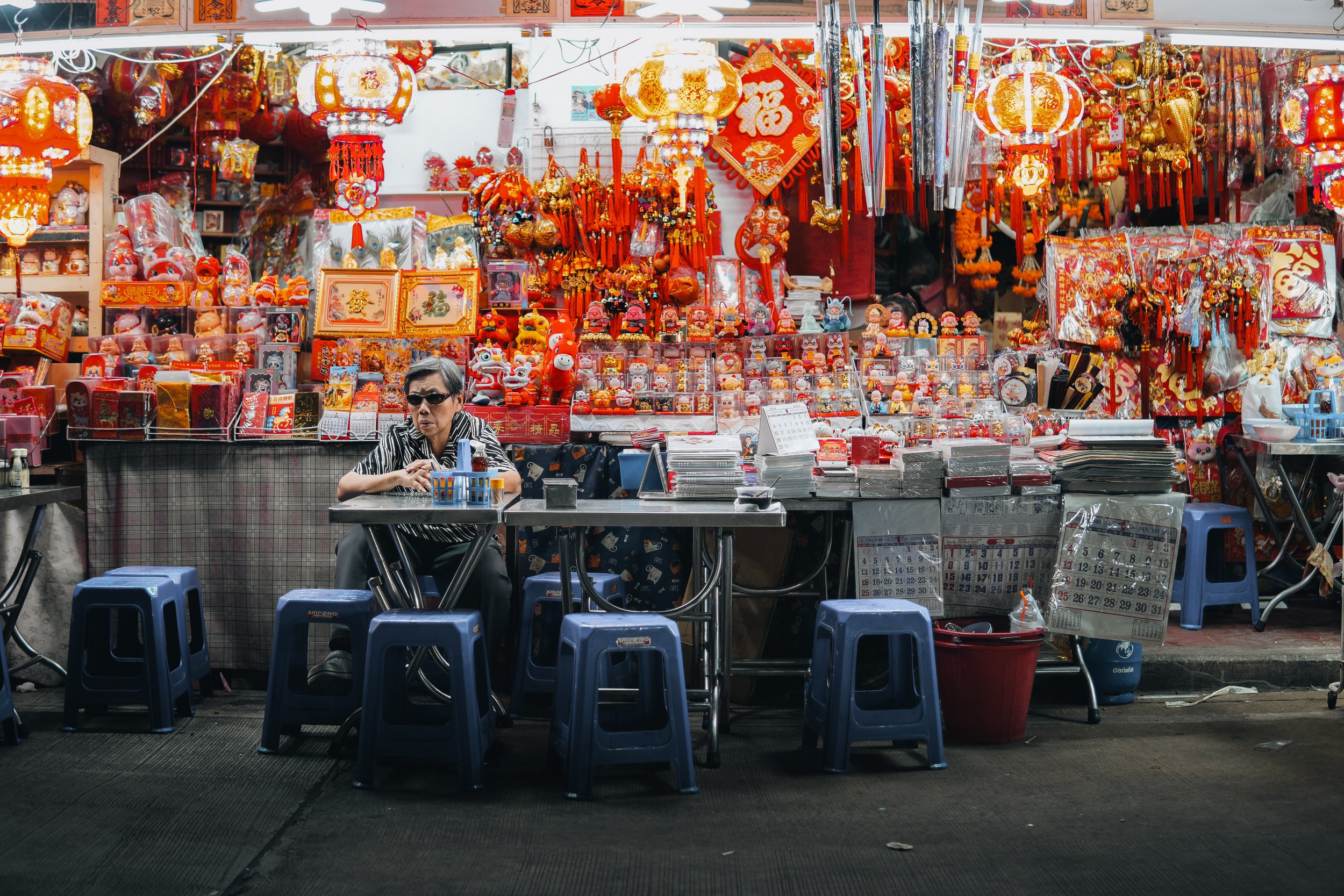 A street vendor's stall decorated with red and gold Chinese New Year decorations, lanterns, stuffed animals, and celebratory items, with a woman sitting at the counter.