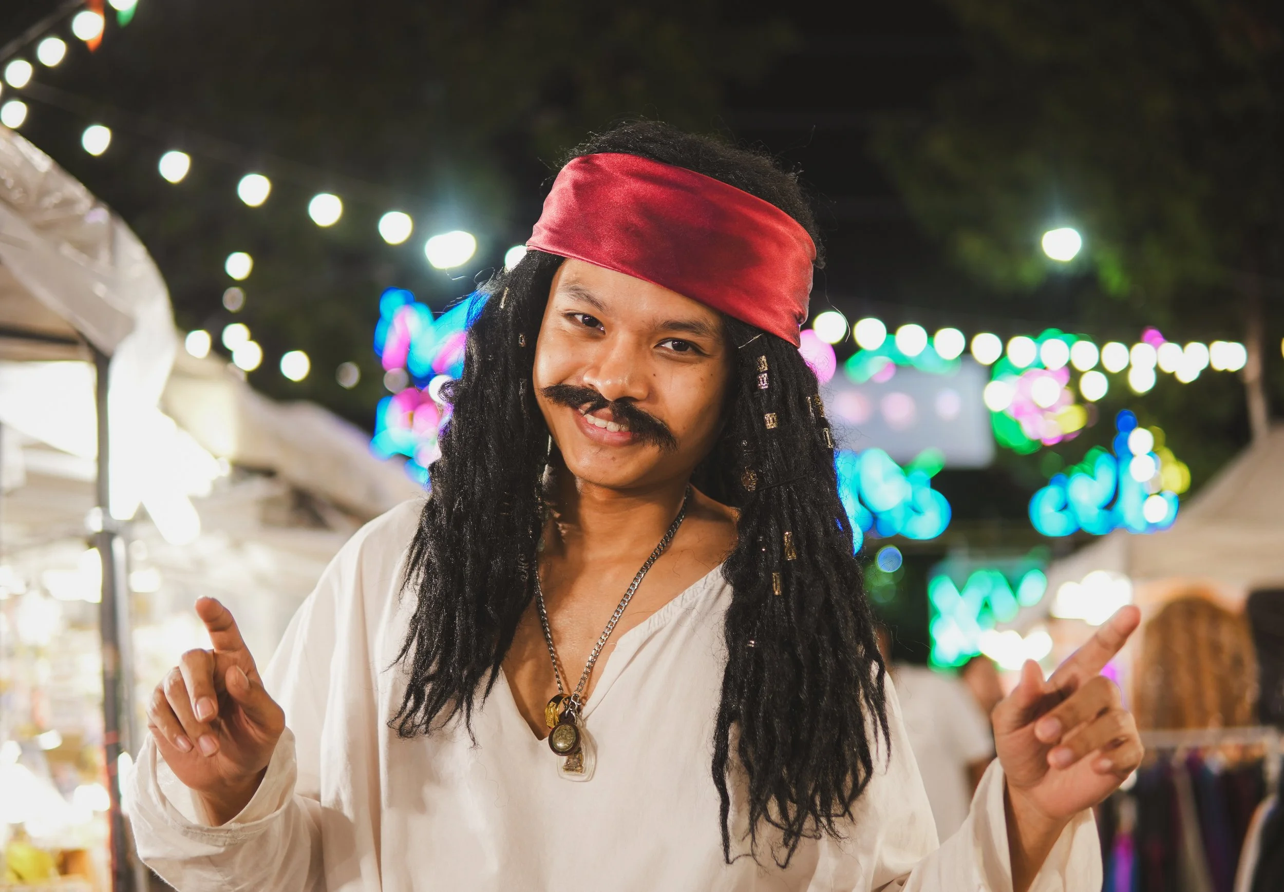 Person dressed as a pirate in a costume at a nighttime outdoor event or festival with colorful lights in the background.