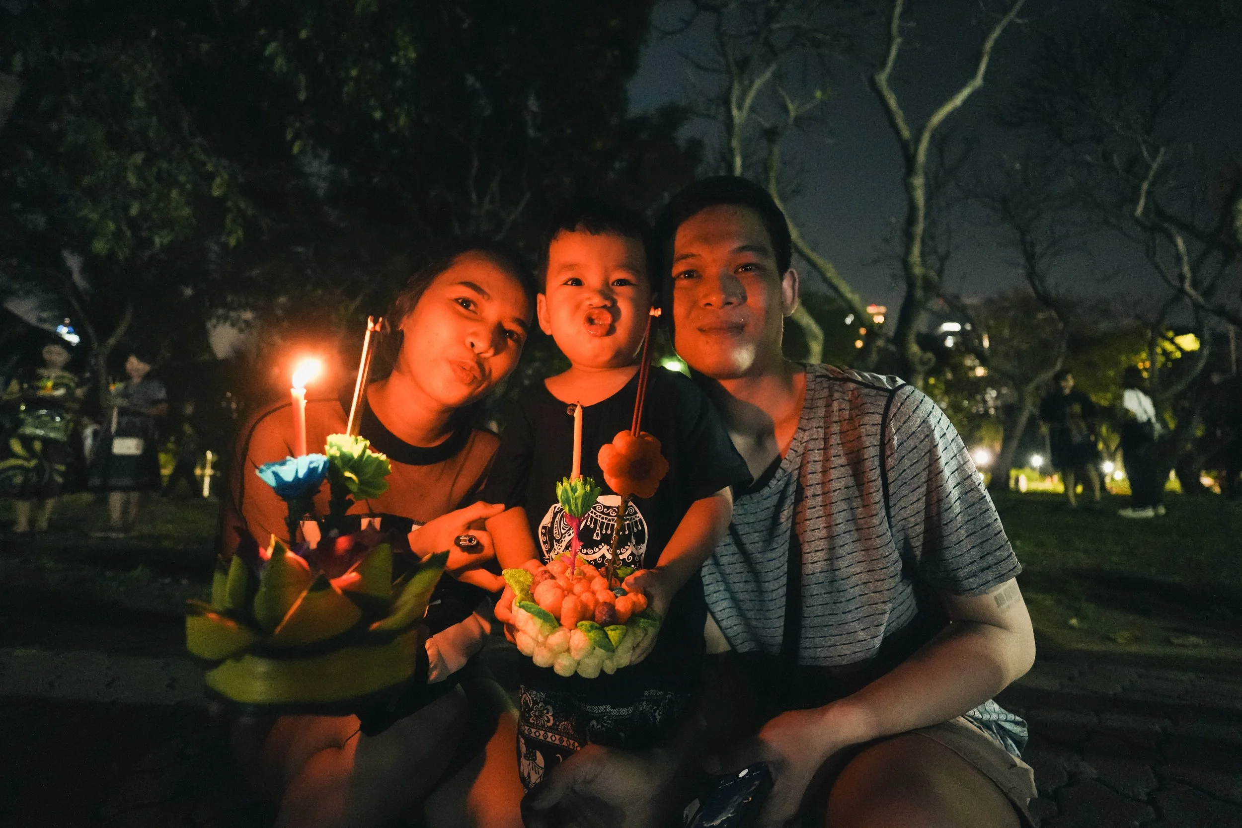 A family of three celebrating at night, holding a birthday cake and colorful candles, with trees and other people in the background.