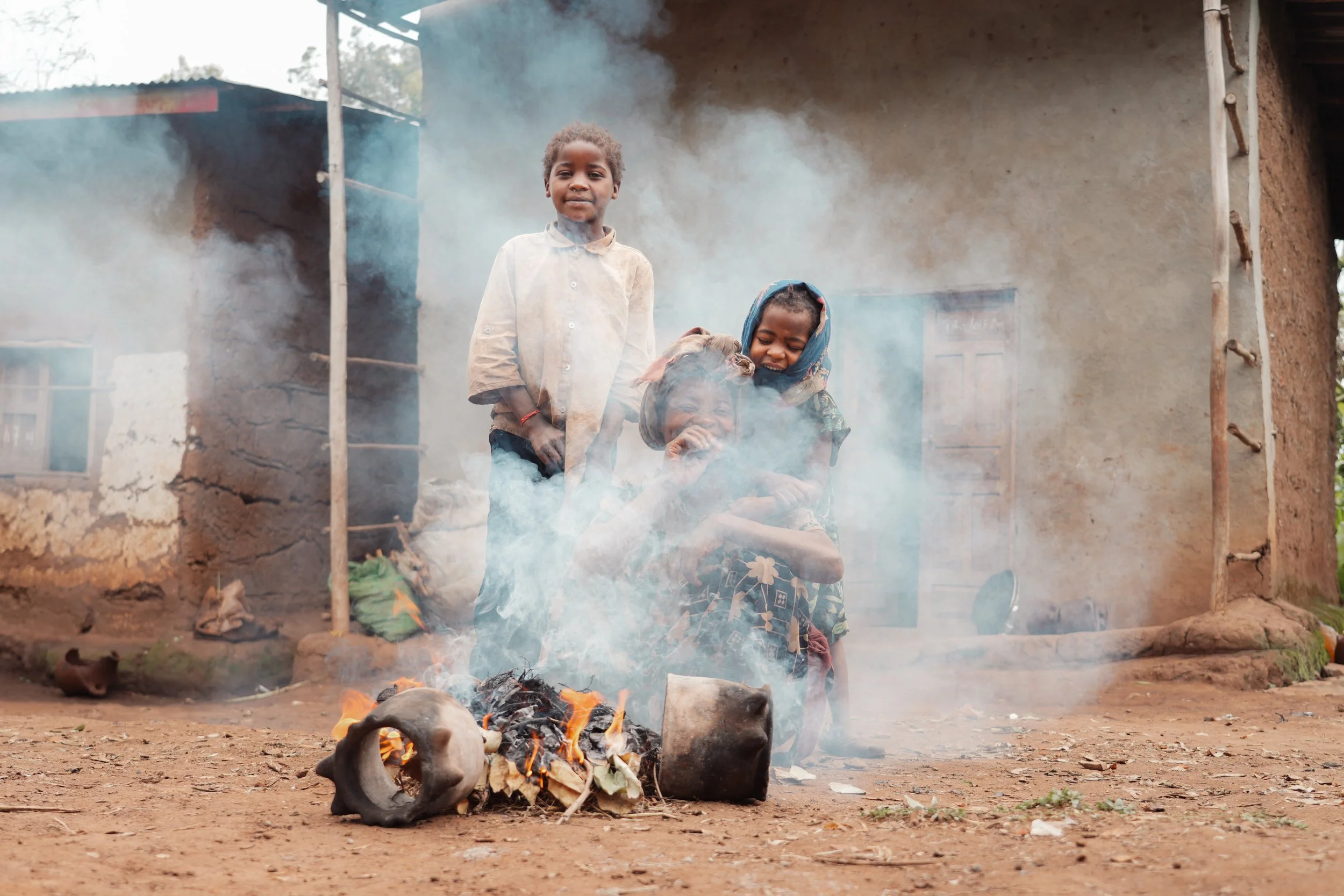 Three children standing near a burning fire with a skull, in front of a mud house. The scene appears rustic and outdoors.