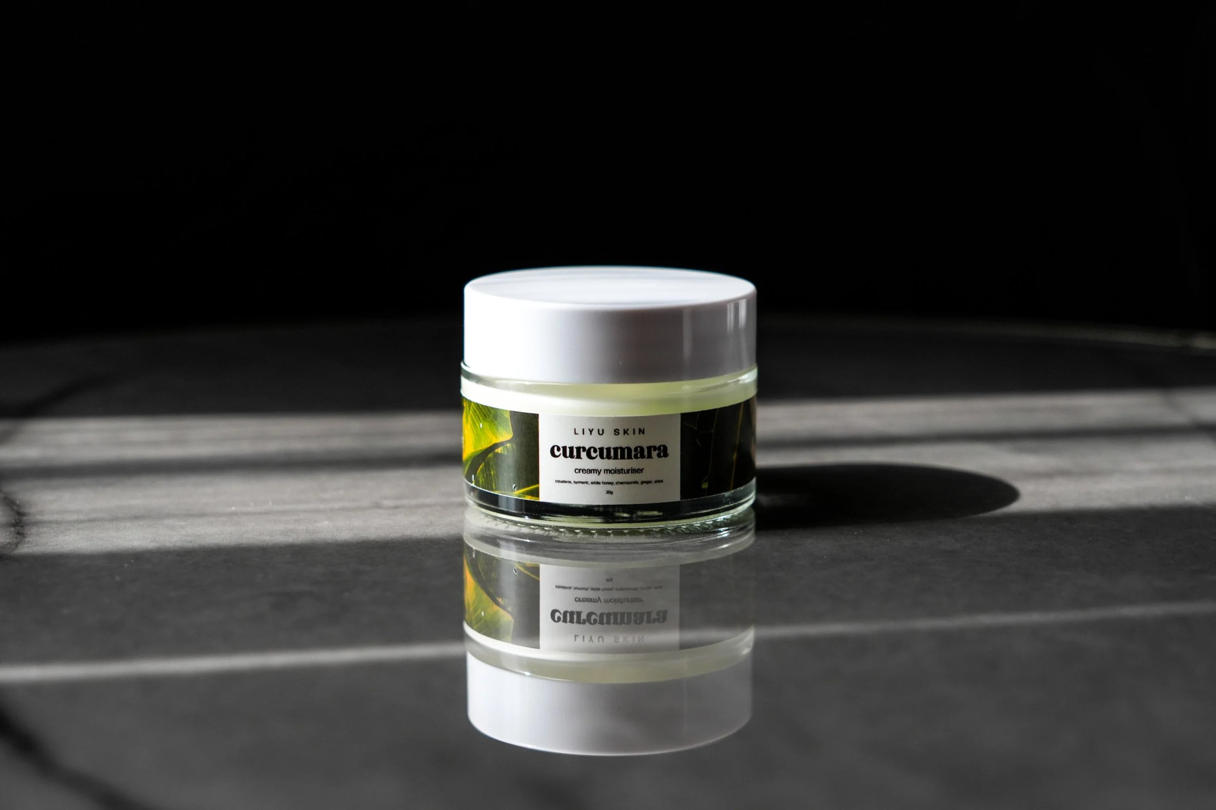 A jar of LIYU SKIN curcumara creamy moisturizer on a dark surface with a reflection, illuminated by light coming from the left.