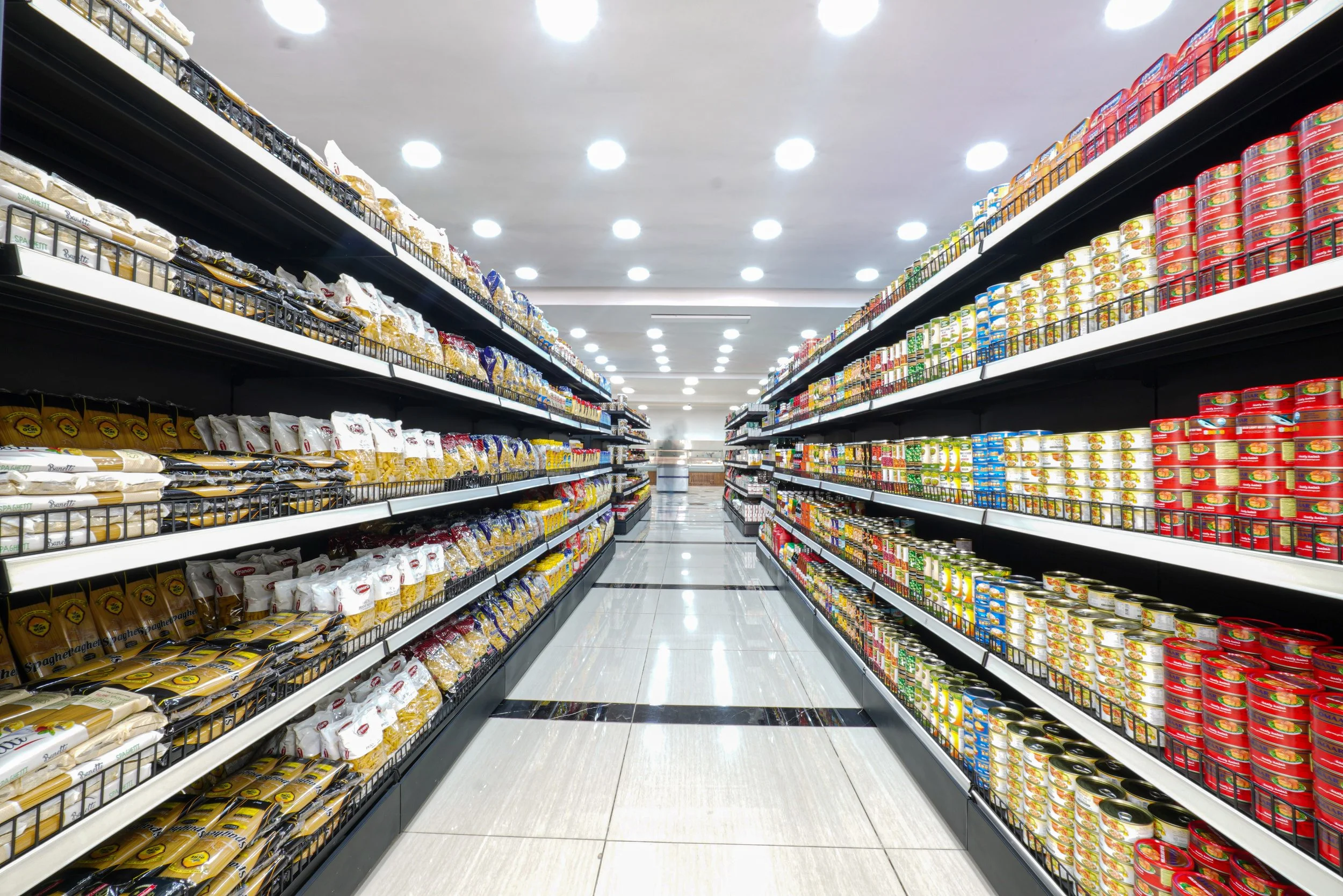 A supermarket aisle with shelves stocked with packaged pasta and canned goods, lit by bright ceiling lights.