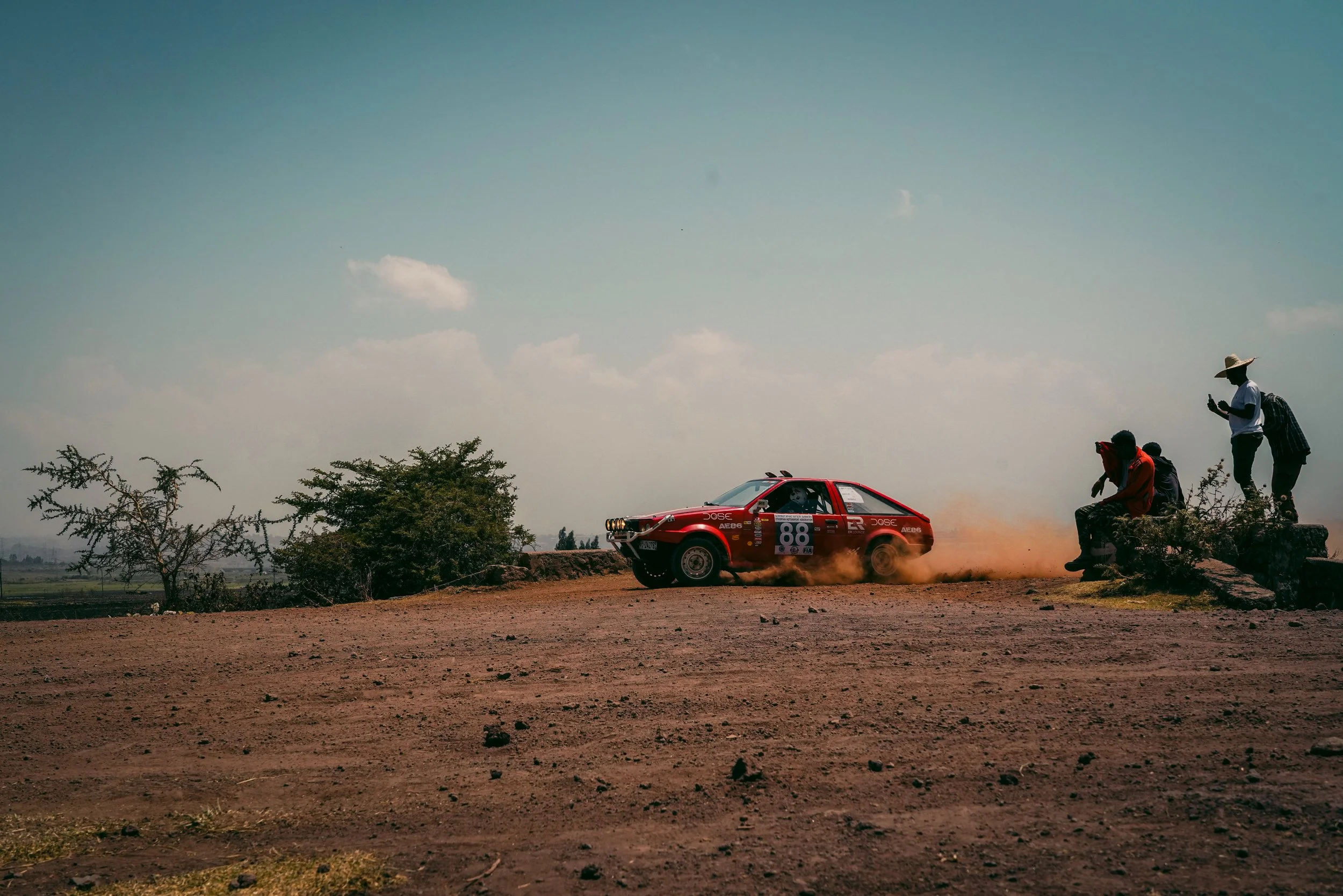 A red rally car with the number 88 on its side is speeding on a dirt road, kicking up dust as it navigates a turn. Three spectators are sitting and standing on a small stone platform watching the race, with one person wearing a large straw hat and ch
