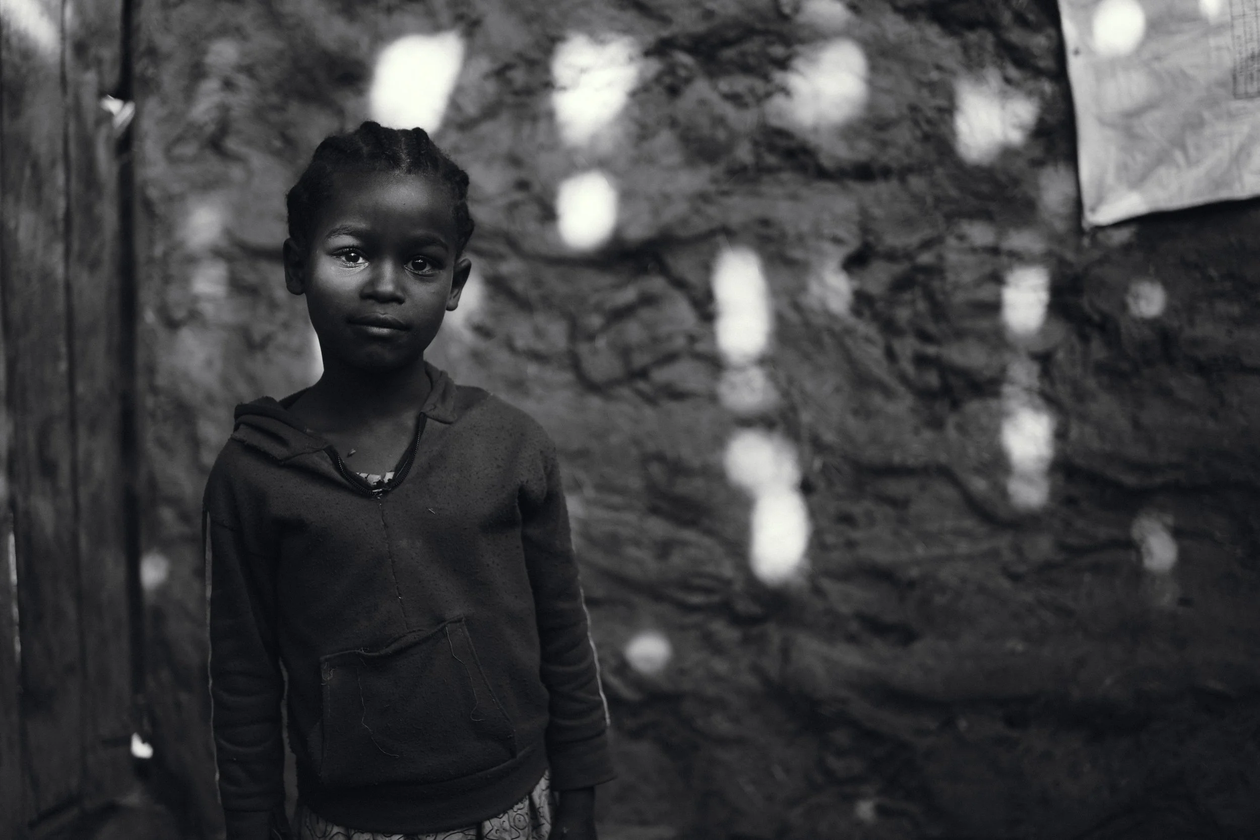A young girl with braided hair standing inside a rustic, possibly mud-walled shelter, looking directly at the camera.