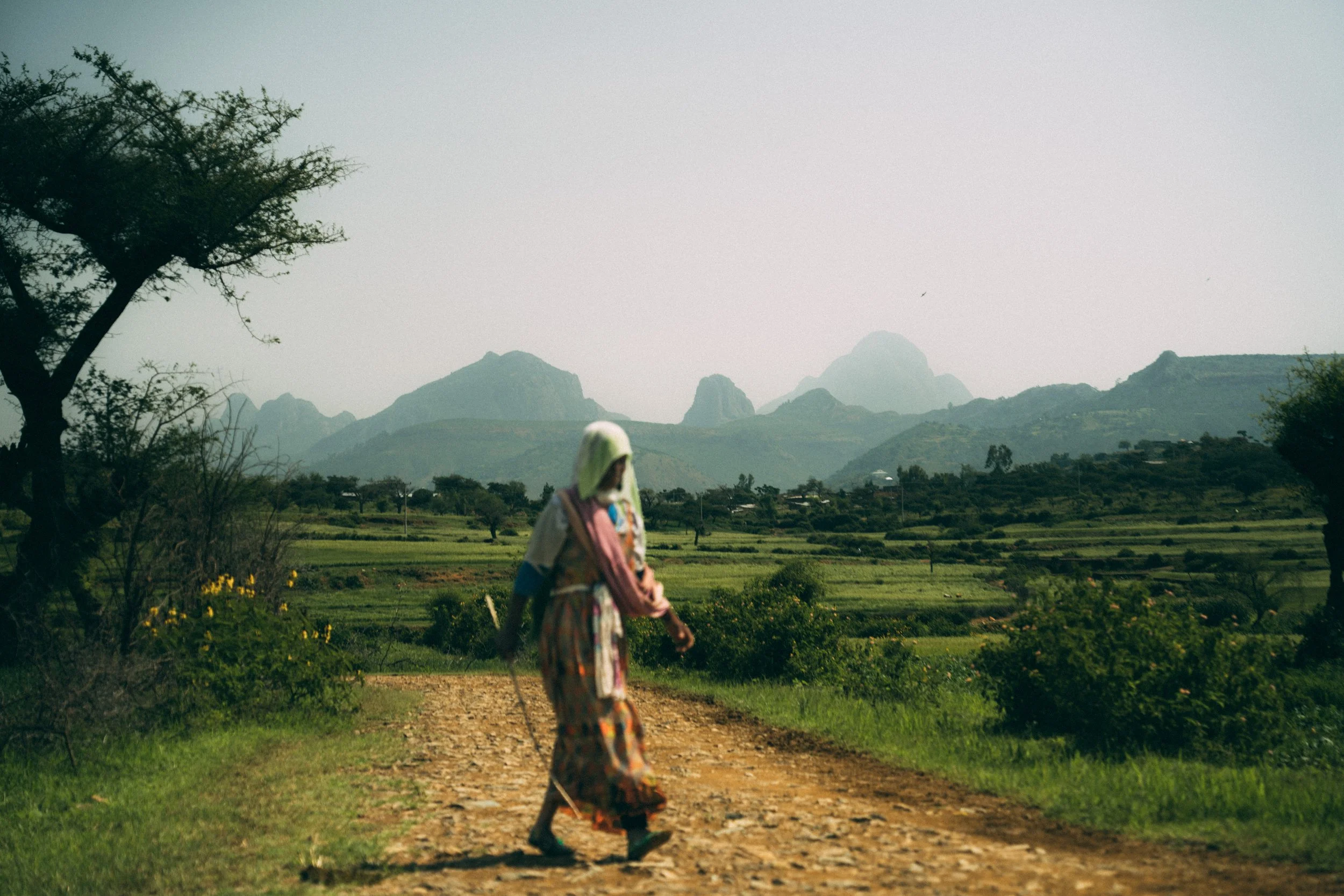 A woman walking on a dirt path through a rural landscape with green fields, trees, and mountains in the background.