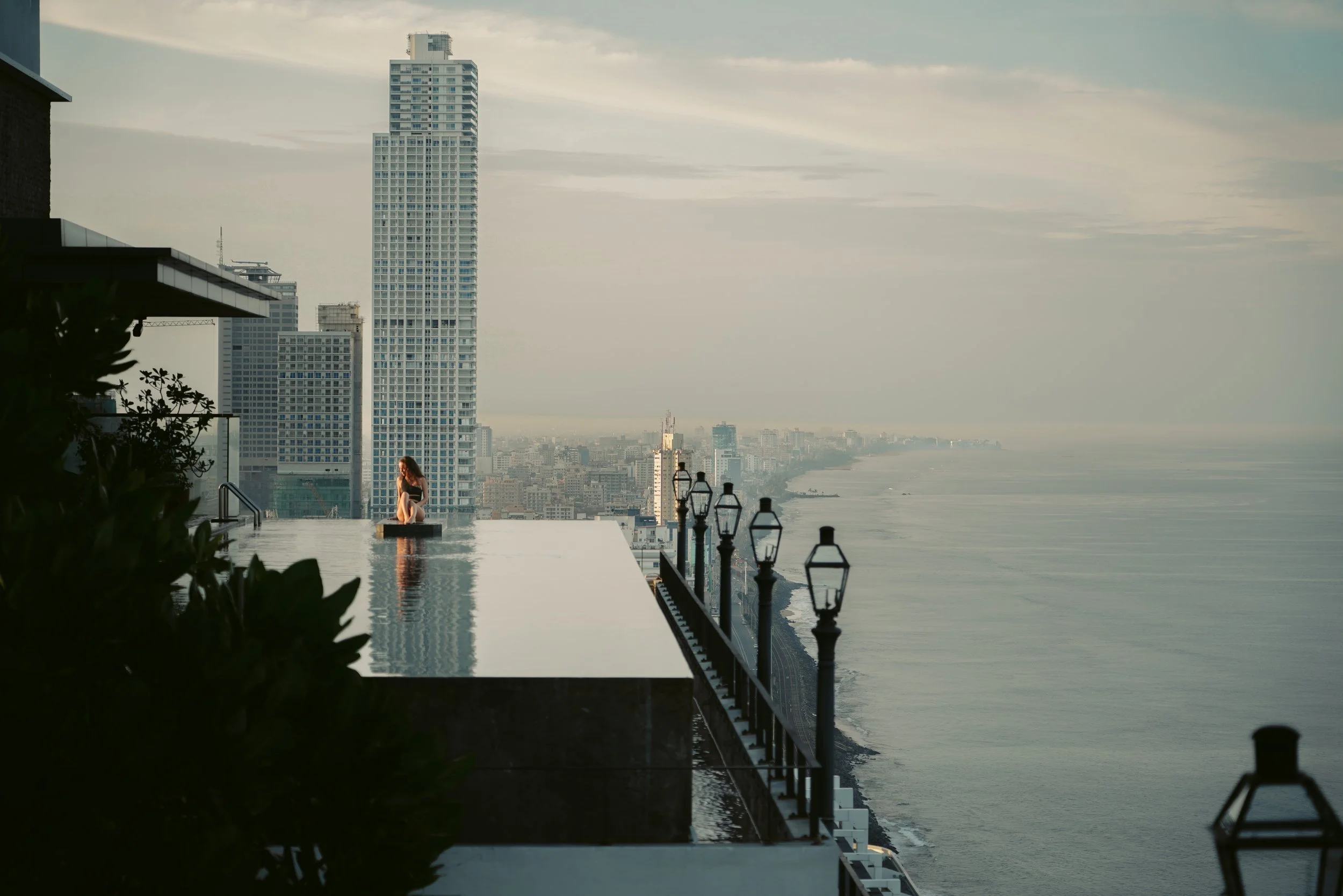 A woman sitting on the edge of an infinity pool on a rooftop with a city skyline and ocean in the background.