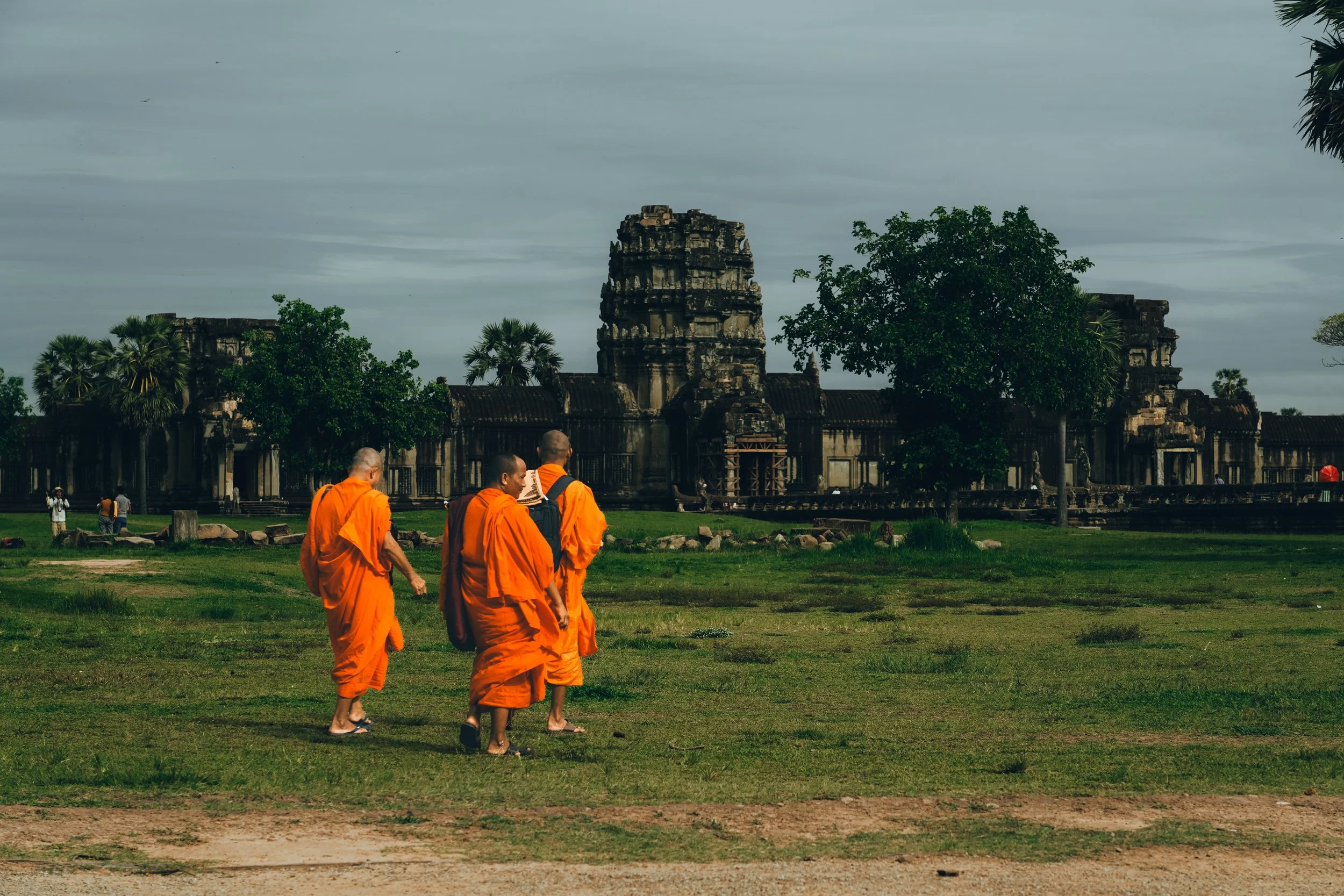 Four monks dressed in orange robes walking across grass near an ancient temple with stone structures and trees.